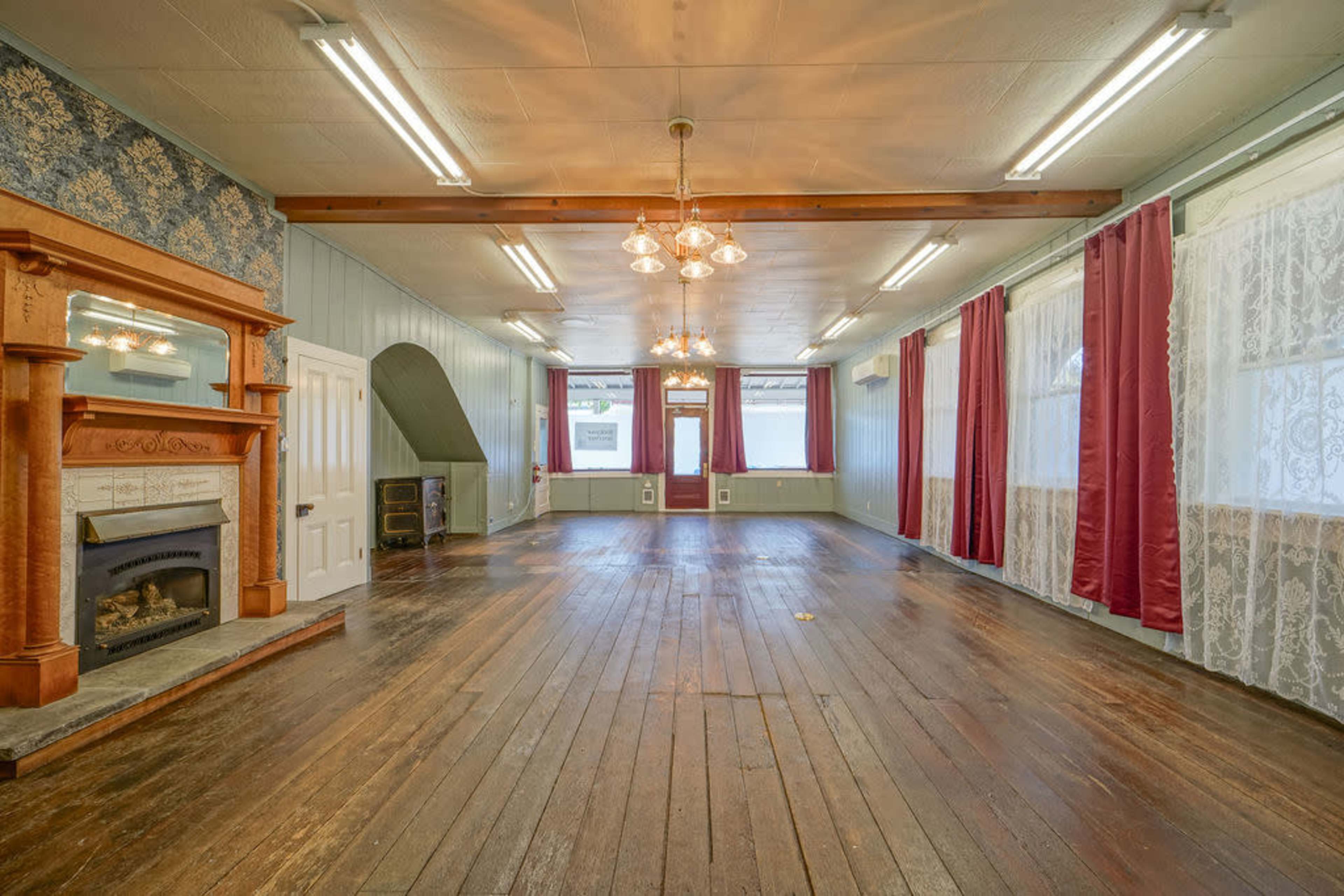 The image shows a spacious room with wooden floors, large windows draped with red curtains, a decorative fireplace, and several ceiling lights.