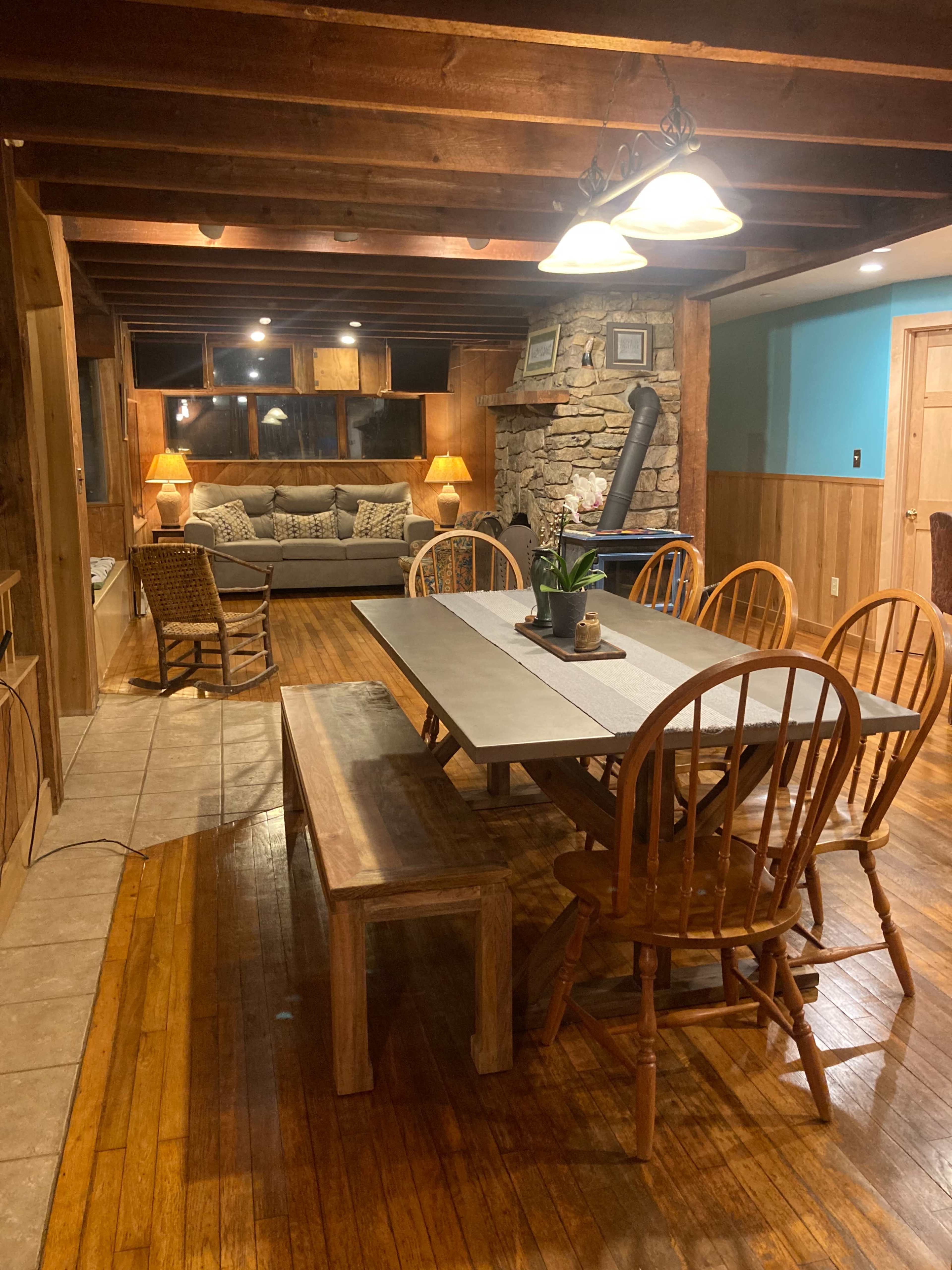 A wooden dining area with a long table and chairs, adjacent to a cozy living space featuring a couch and a stone fireplace.