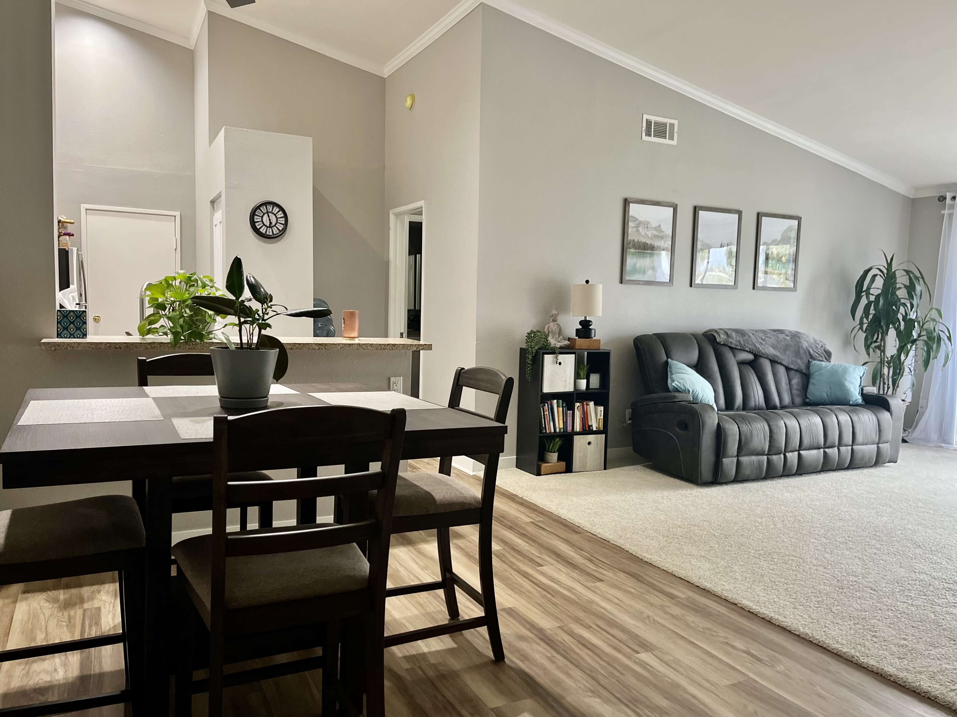 The image shows a spacious living area with a dining table, a gray sofa, and a large window, featuring neutral-colored walls and wood flooring.