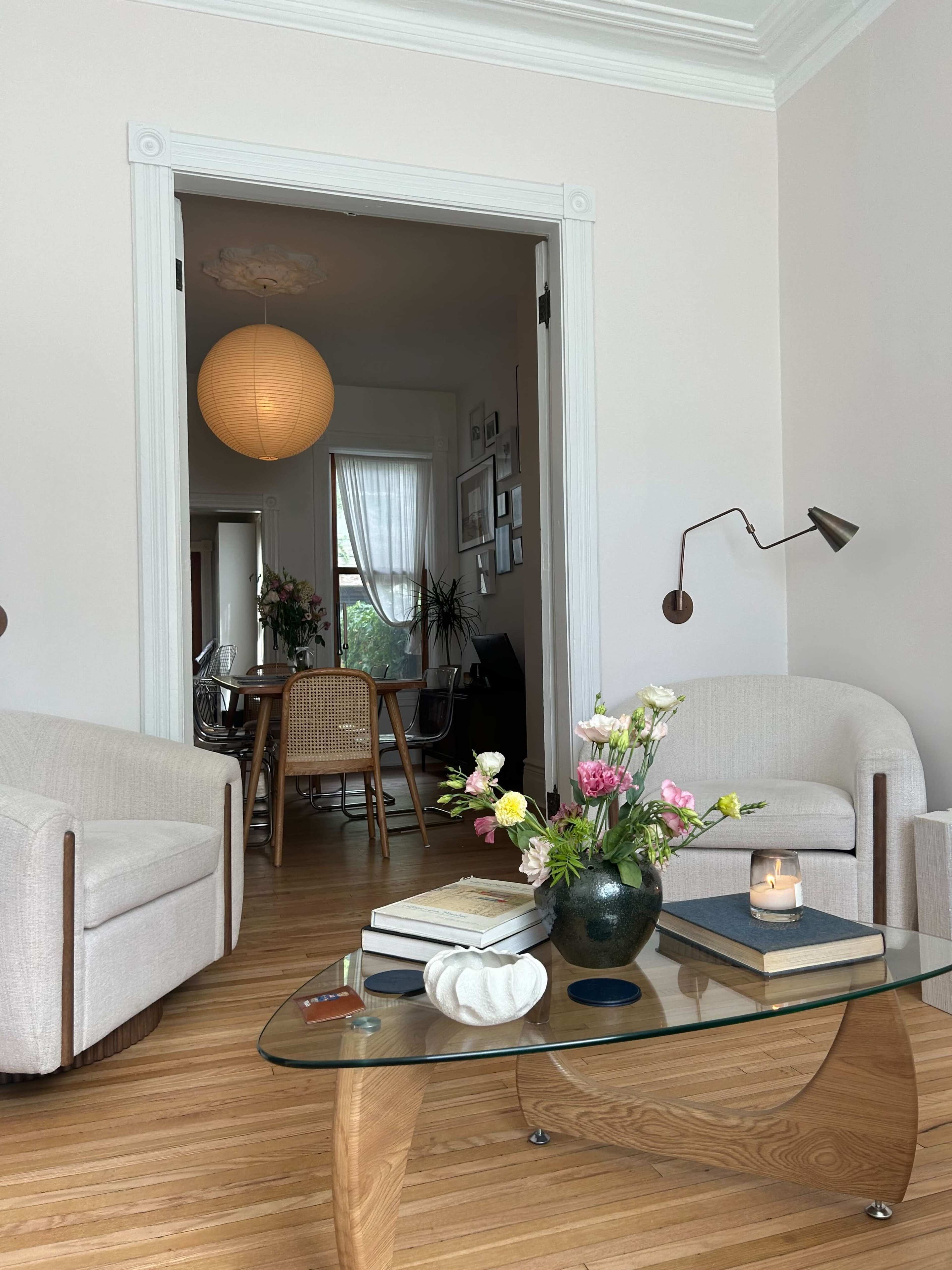 A cozy living room features two light-colored armchairs, a glass coffee table with a floral arrangement, and an open doorway leading to another room.
