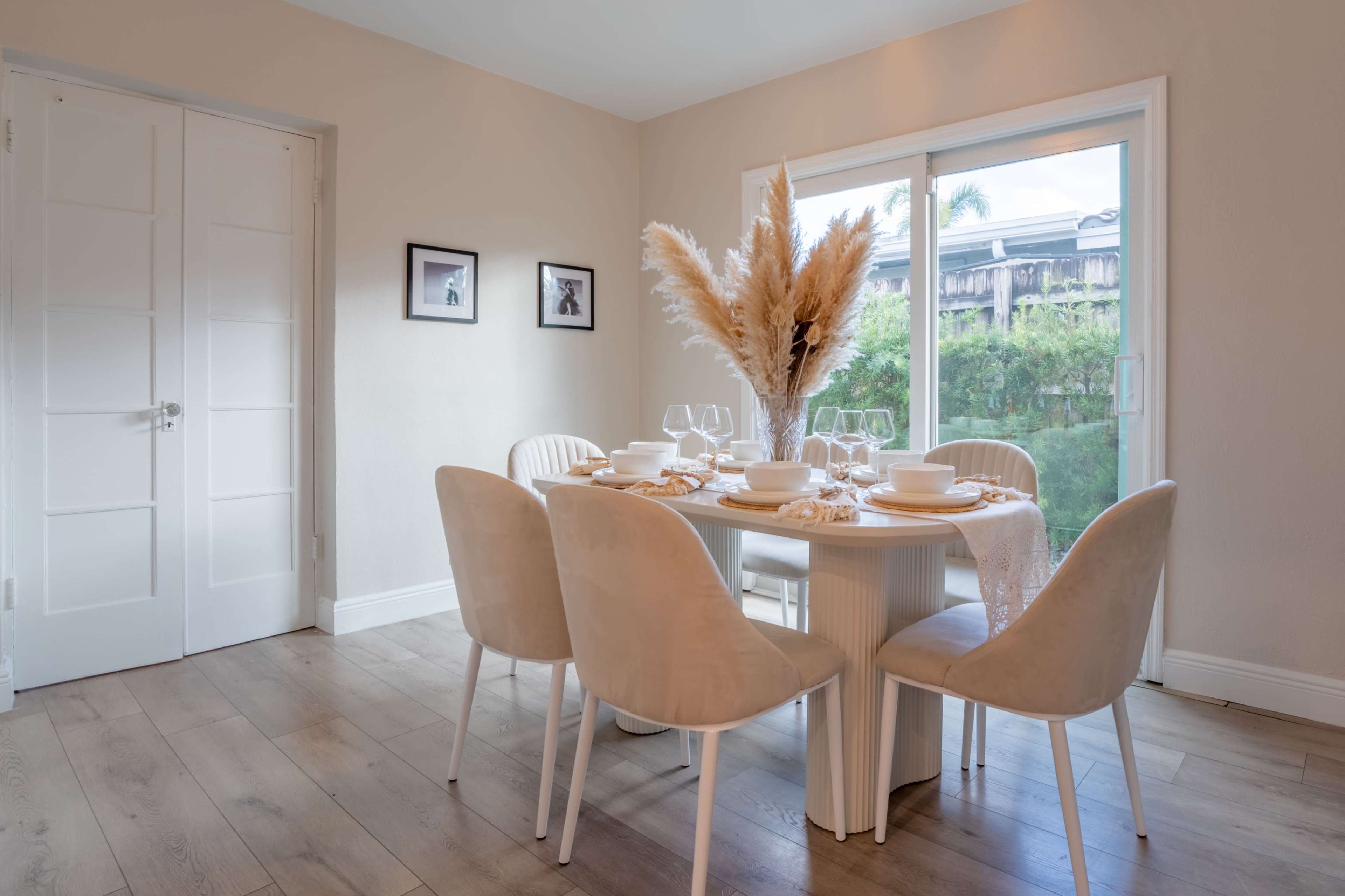 A dining room features a round table set with dinnerware and surrounded by six upholstered chairs, with large windows providing natural light and a view of greenery outside.