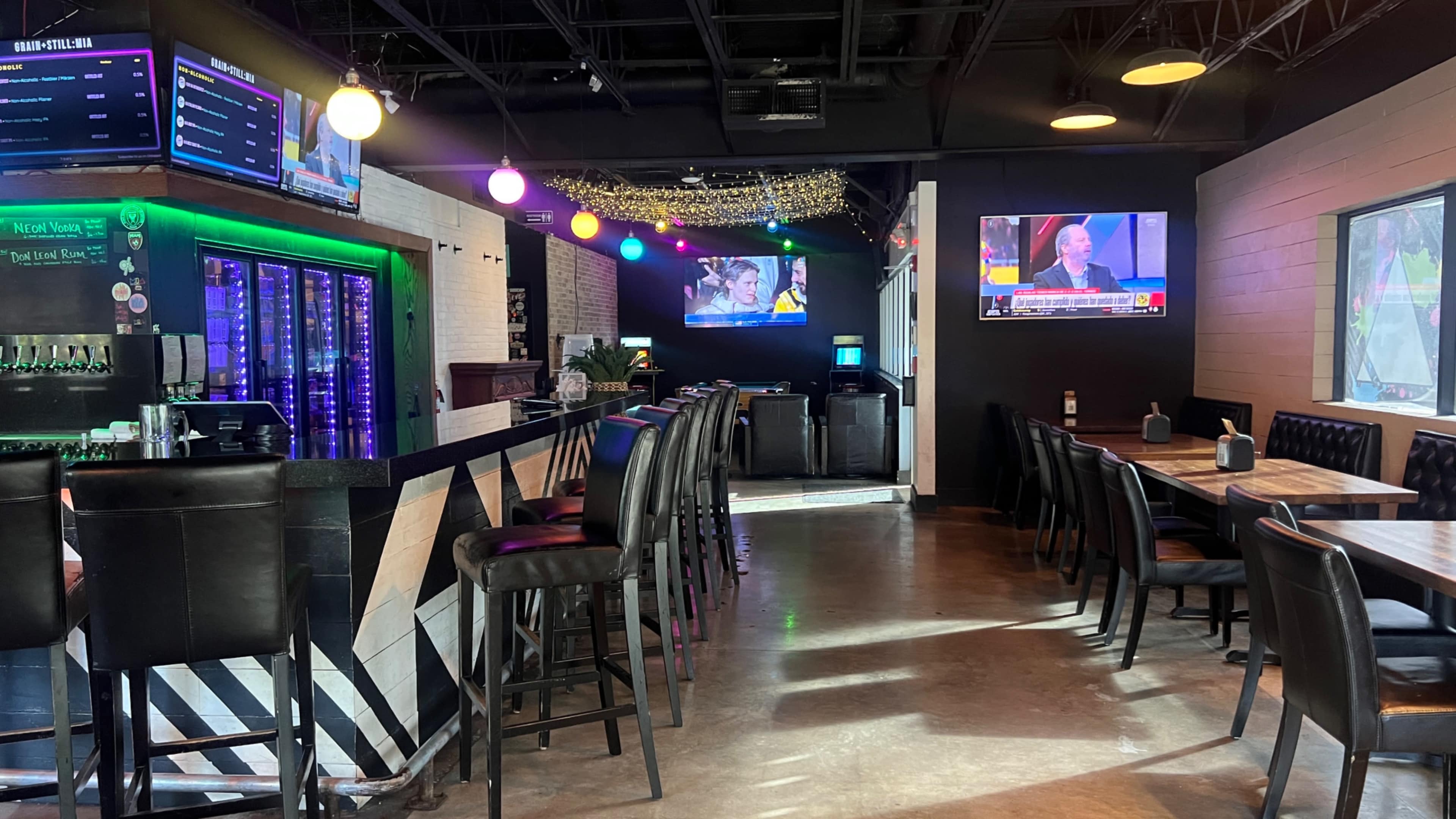 The image shows the interior of a bar or restaurant with a long bar, several tables, and multiple screens displaying sports and news.