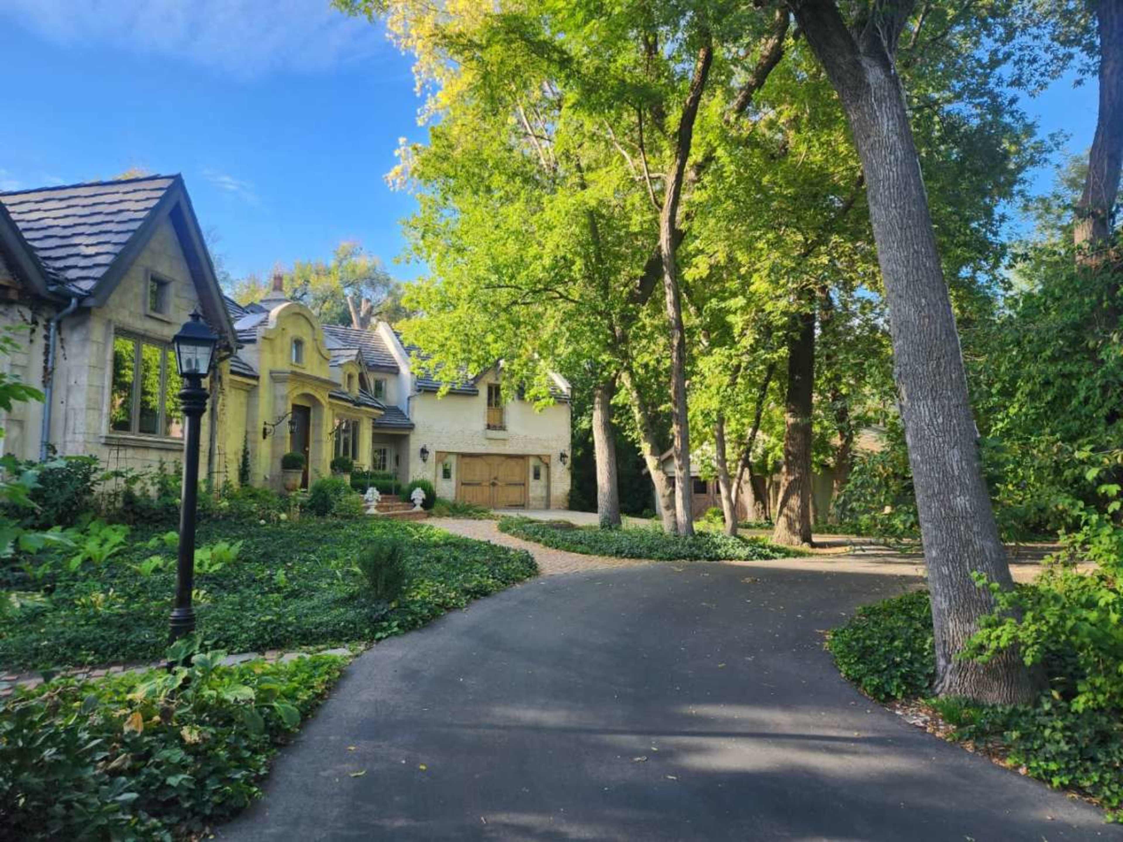 A winding driveway leads to a large, stately home surrounded by lush greenery and tall trees on a clear day.