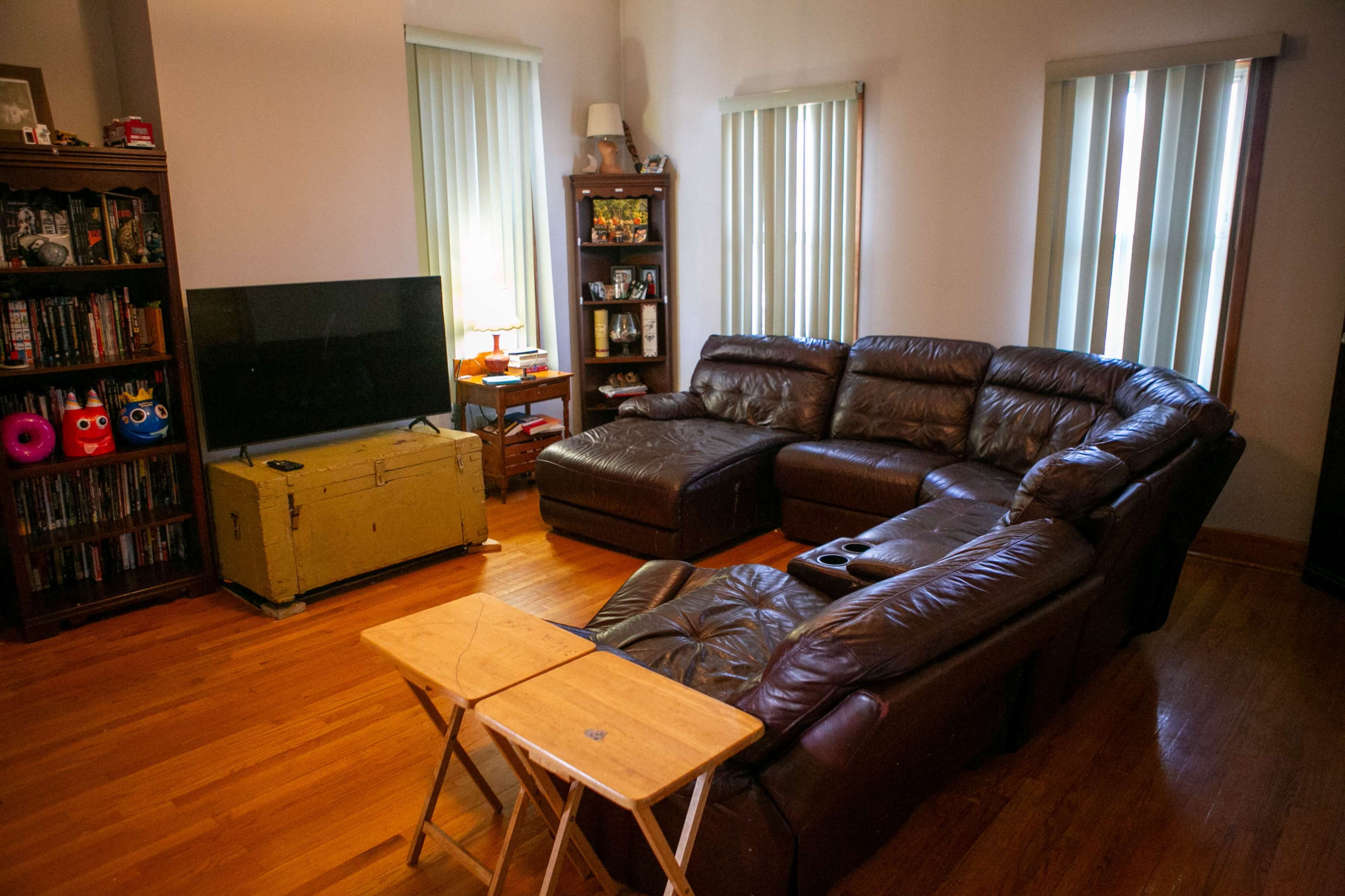 The living room features a brown leather sectional couch, a wooden coffee table, and a vintage chest, with windows dressed in light green blinds, and a television against one wall.