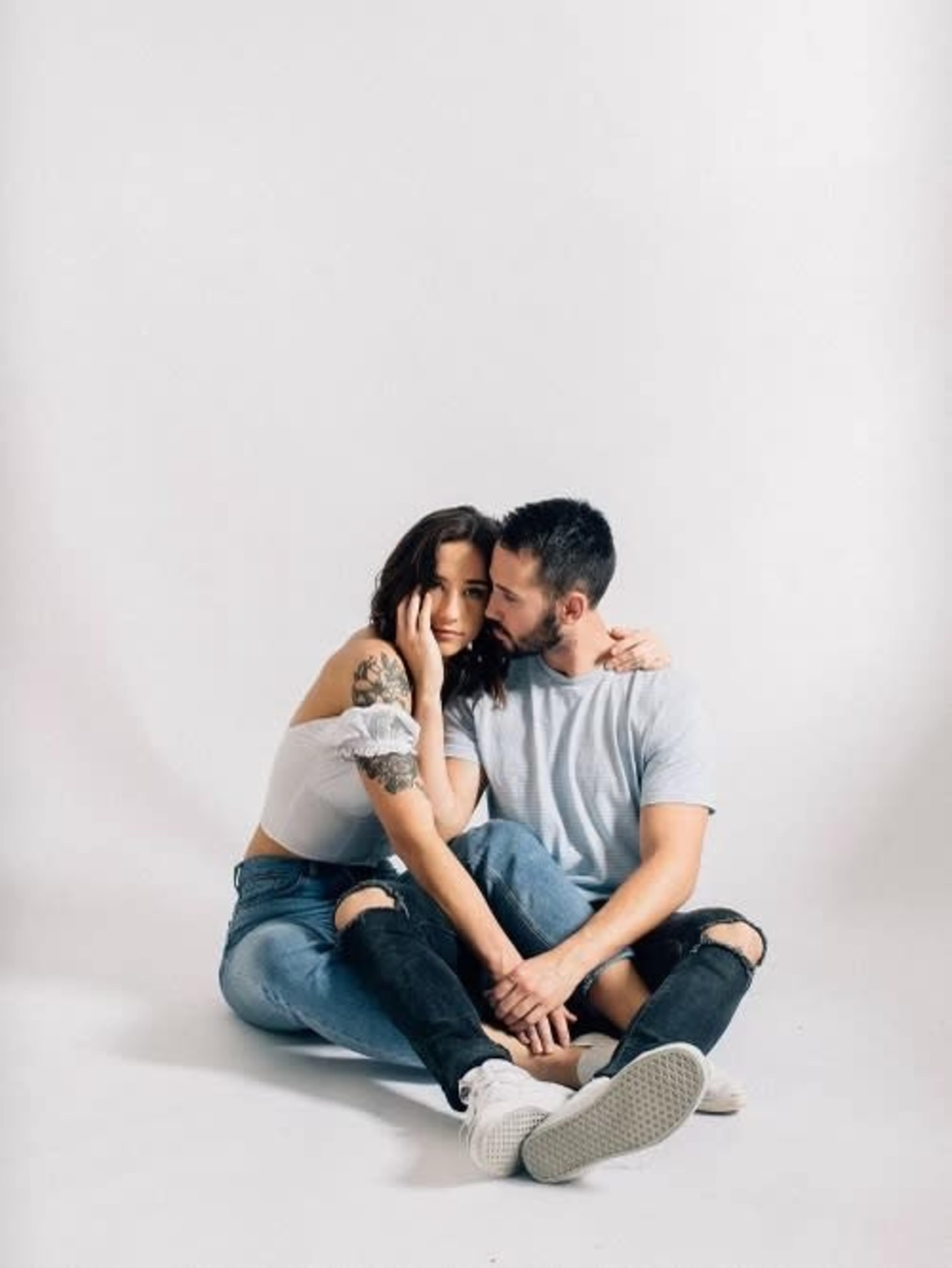 A couple sits on a white floor, with one partner leaning their head against the other's shoulder.
