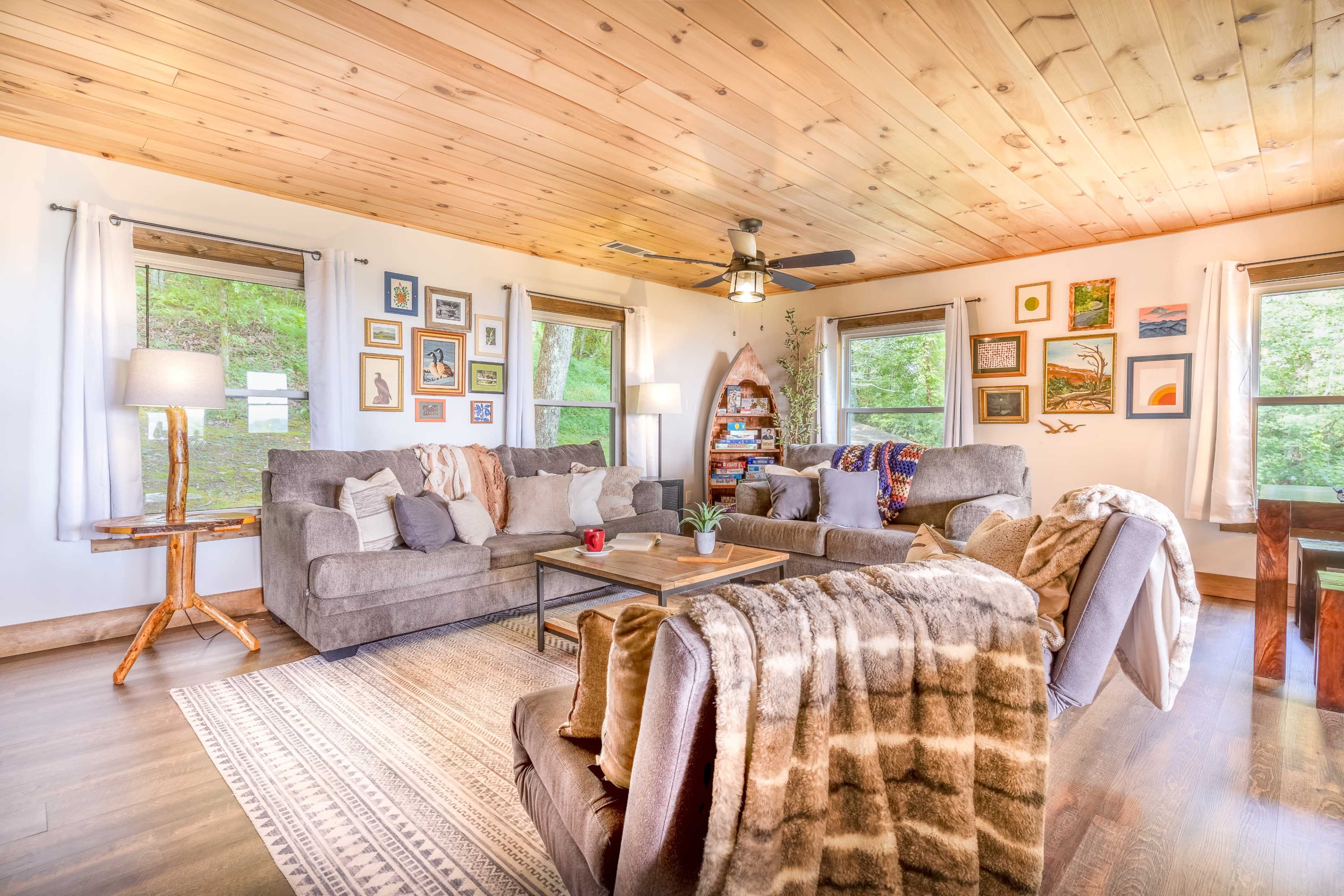 A cozy living room with a wooden ceiling, two large sofas, a coffee table, and various framed pictures on the walls, surrounded by windows letting in natural light.