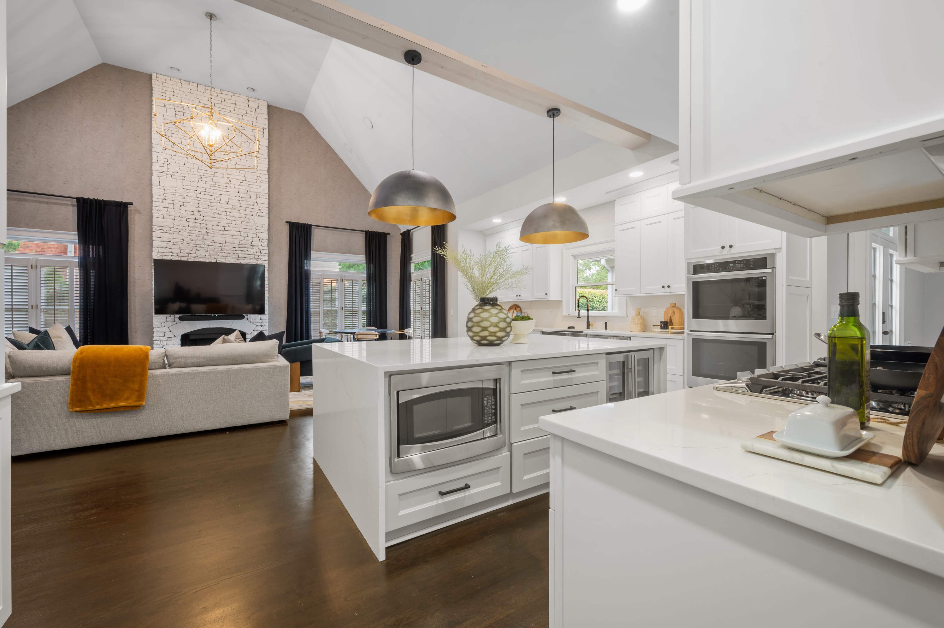 A modern open-concept kitchen and living room featuring white cabinetry, a large island with pendant lighting, and a cozy seating area with a stone fireplace.