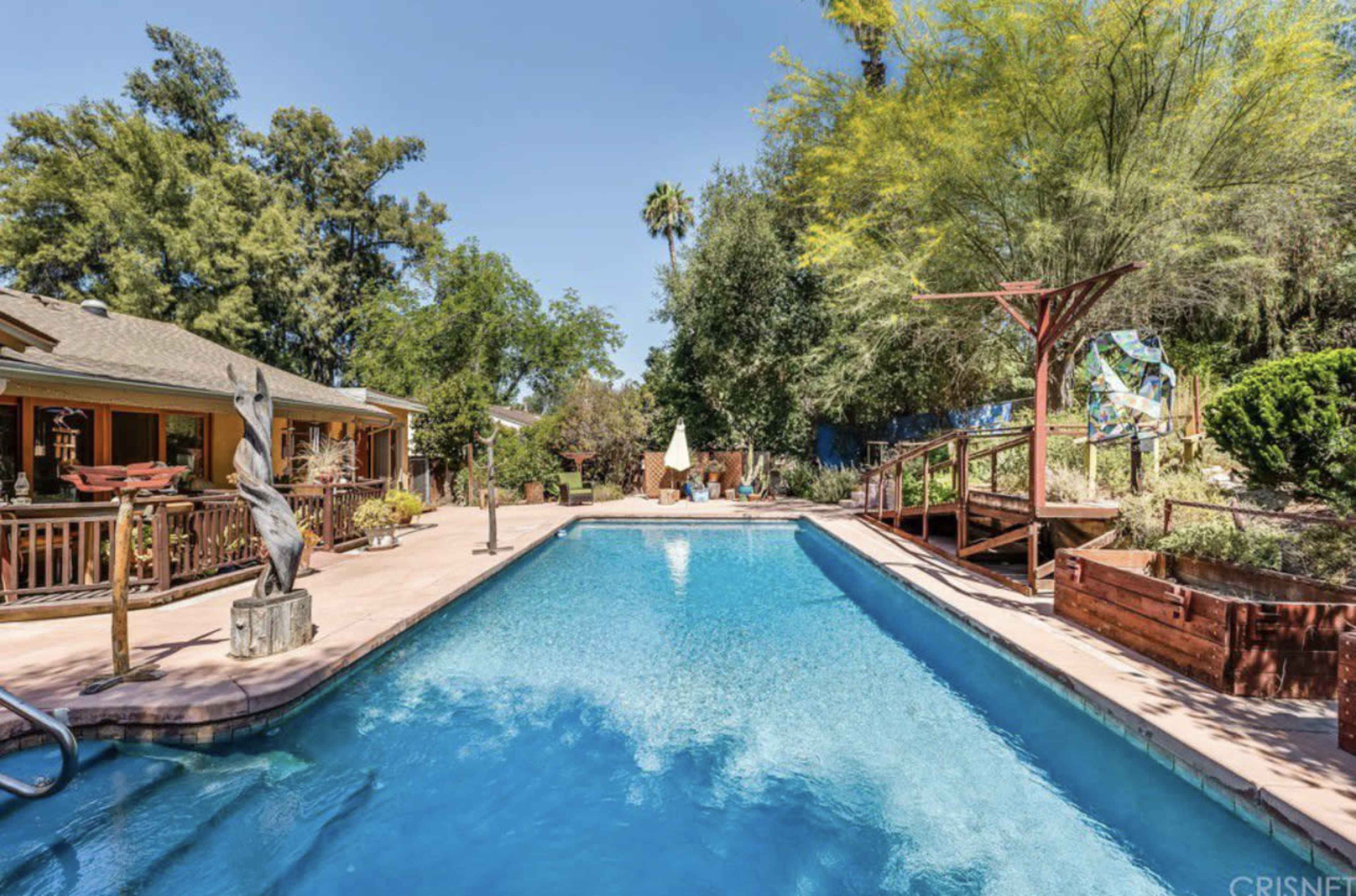 The image shows a backyard swimming pool surrounded by greenery, a wooden deck, and a sculpture.