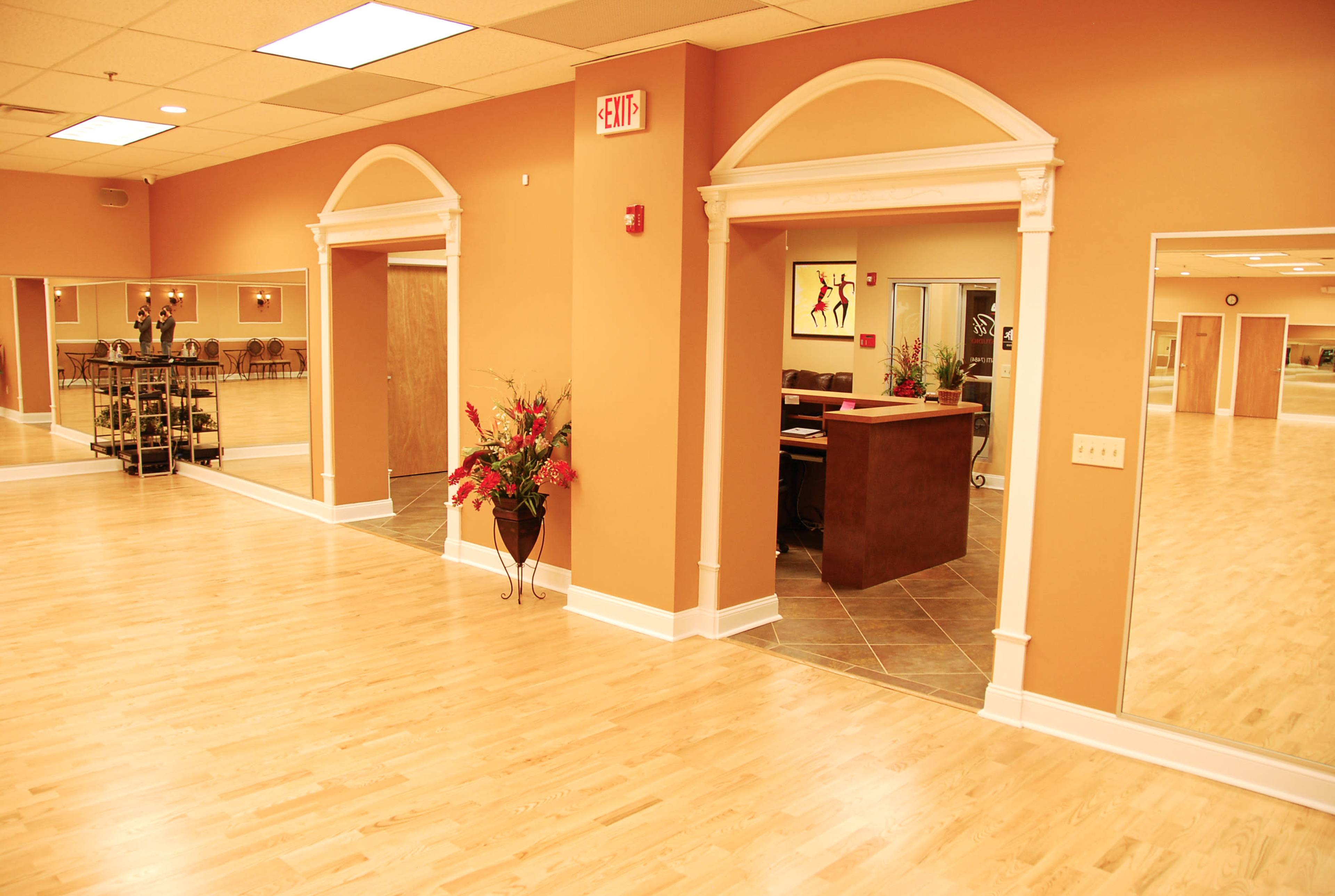 A spacious dance studio with wooden floors, two arched doorways leading to a reception area, and decorative plants in the corners.