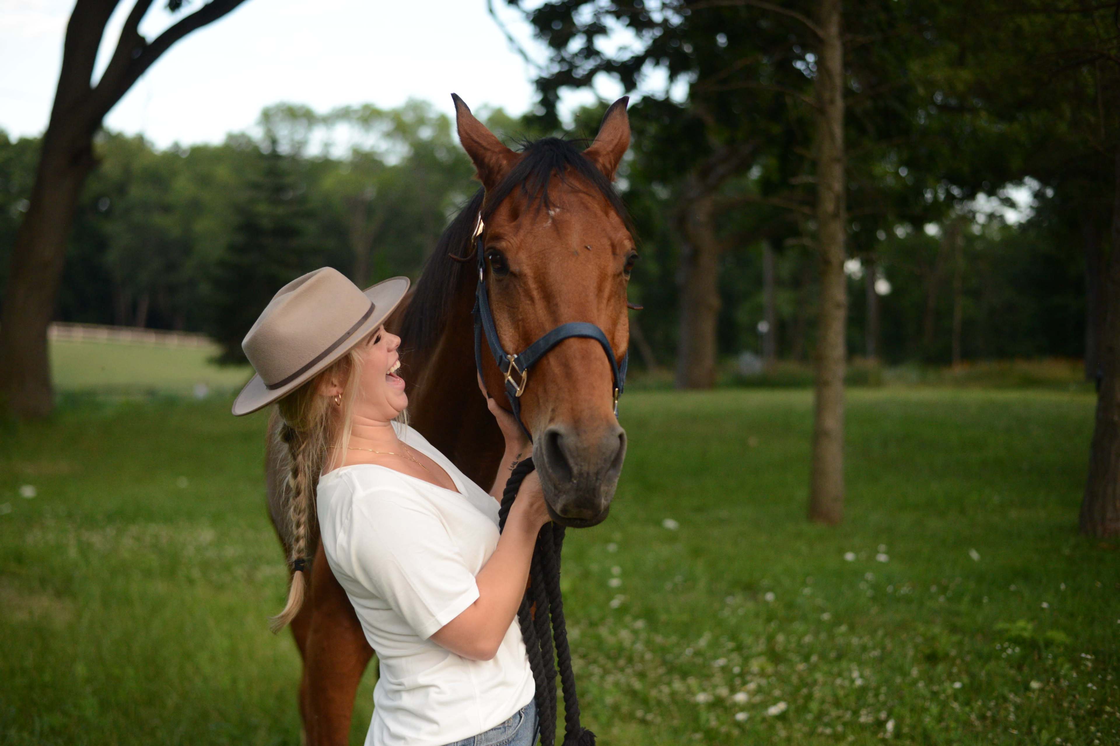 A woman in a white shirt and hat stands beside a brown horse, holding its reins in a grassy field surrounded by trees.