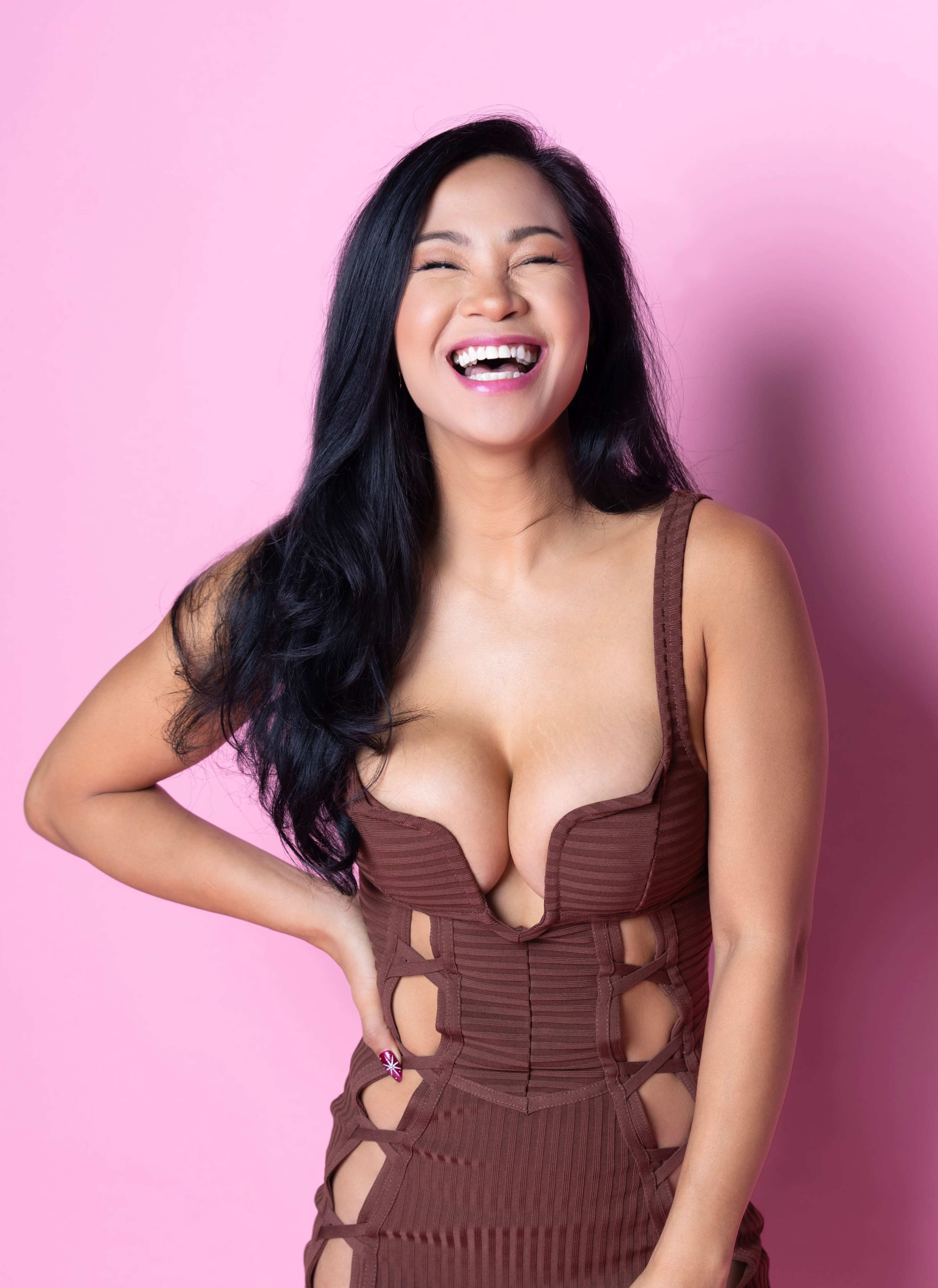 A woman smiles brightly while posing in front of a pink background, wearing a brown, form-fitting dress.