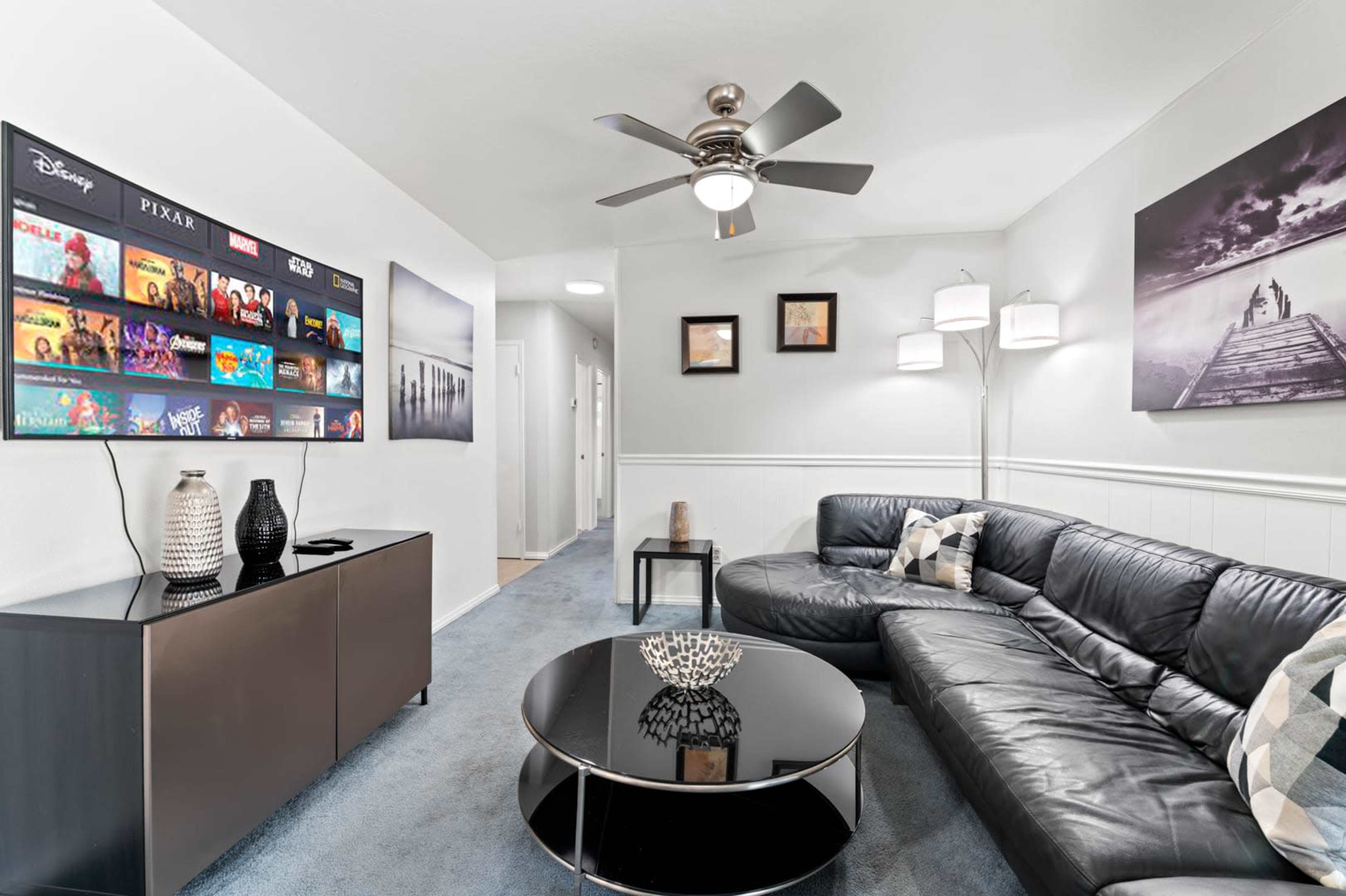 A modern living room features a black leather sectional couch, a round coffee table, and a wall-mounted TV displaying various movie titles.