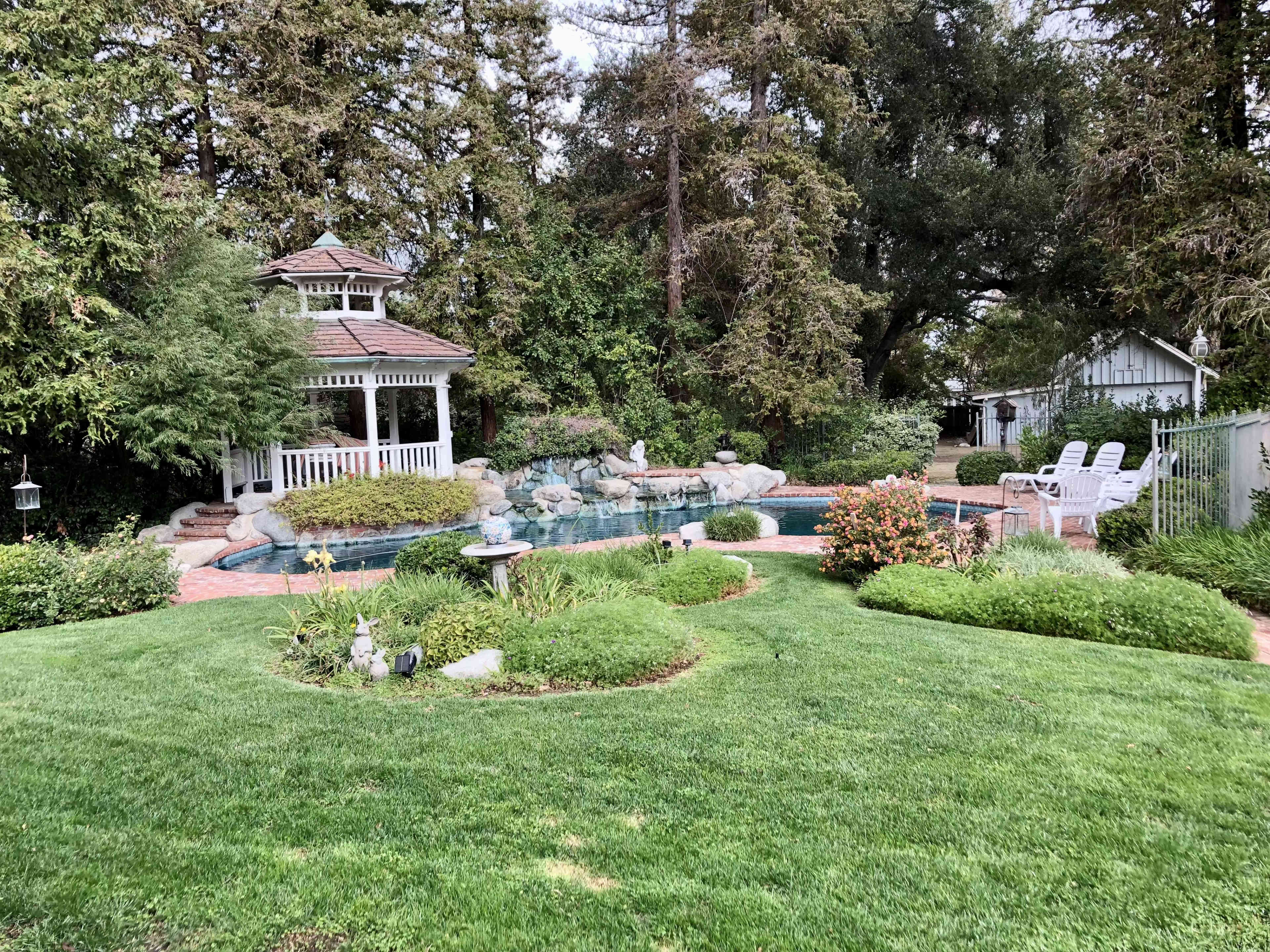 A landscaped backyard featuring a swimming pool, a gazebo, and several garden areas with greenery and flowers.