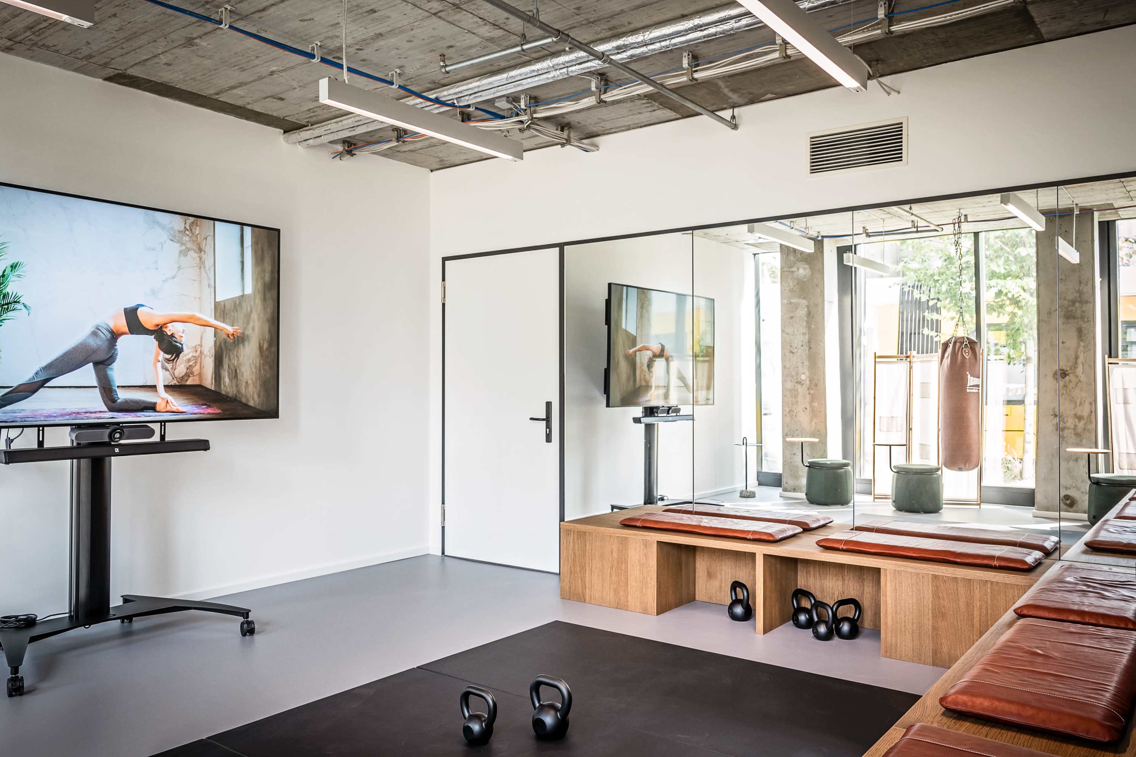 The image shows a modern gym interior featuring exercise mats, kettlebells, a large screen displaying a fitness video, and a mirrored wall with a punching bag.