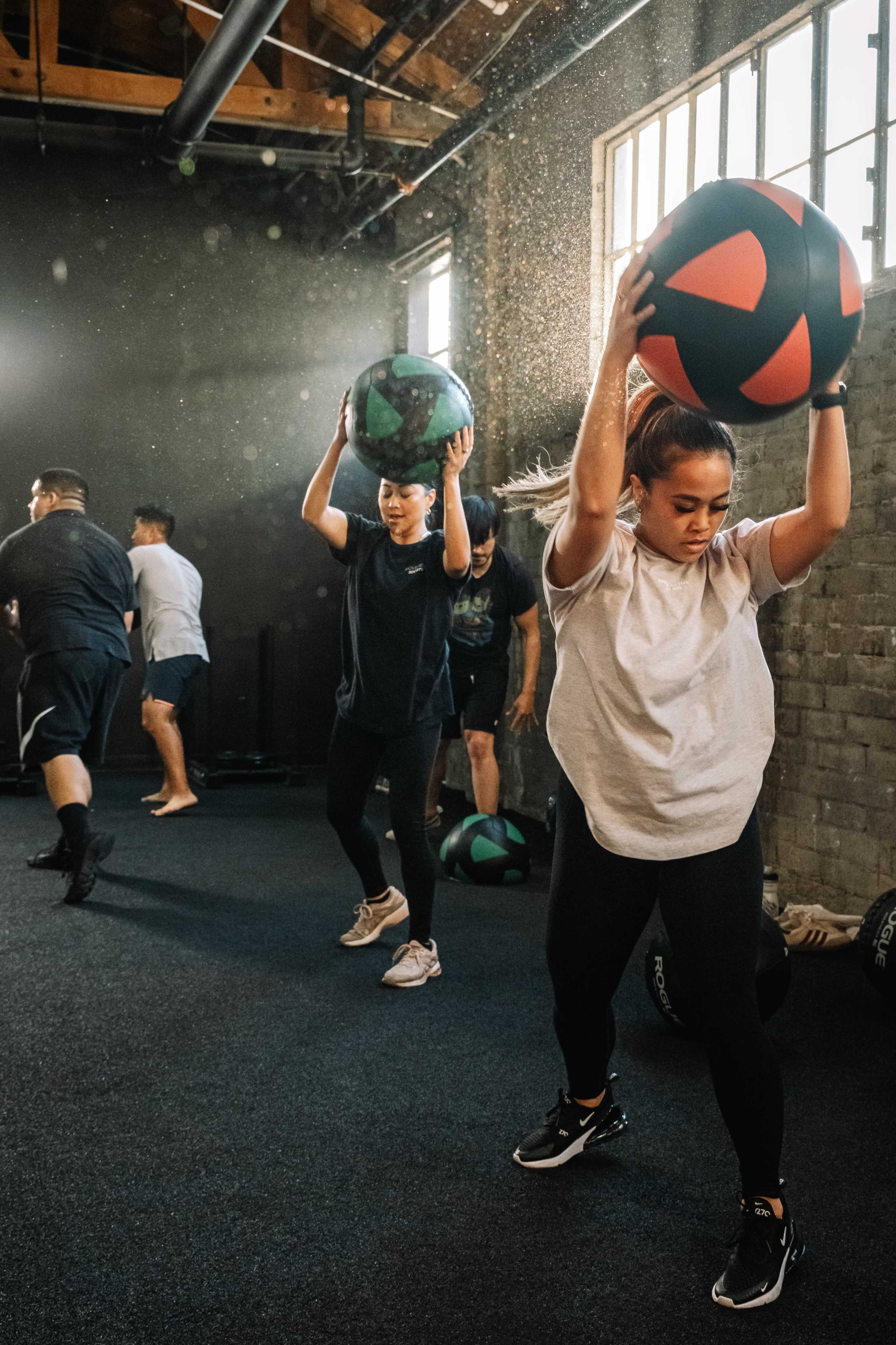 A group of people are exercising with medicine balls in a bright gym with a textured wall.