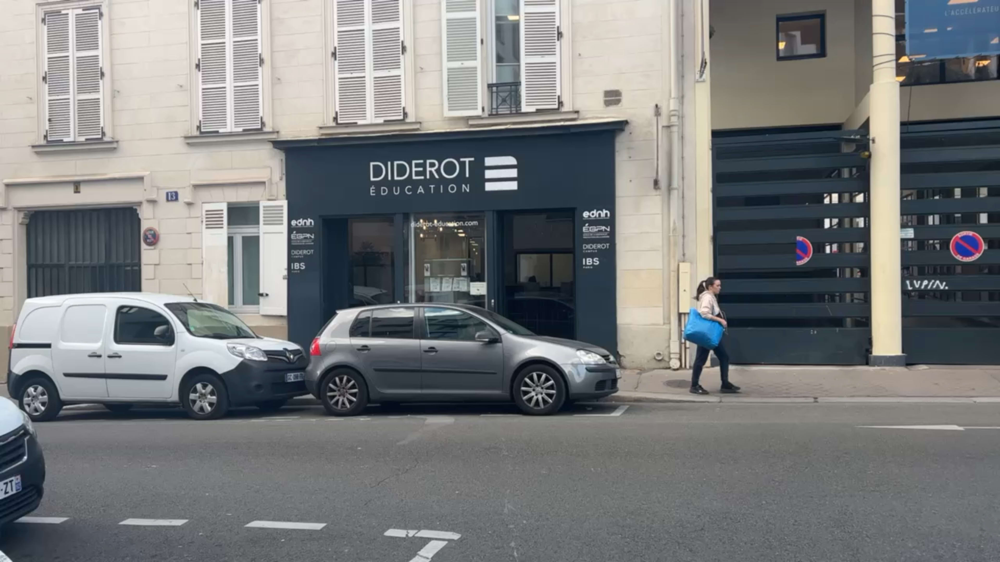 A gray car parked on the street in front of a building with a sign reading "Diderot Education" and a woman walking by carrying a blue bag.