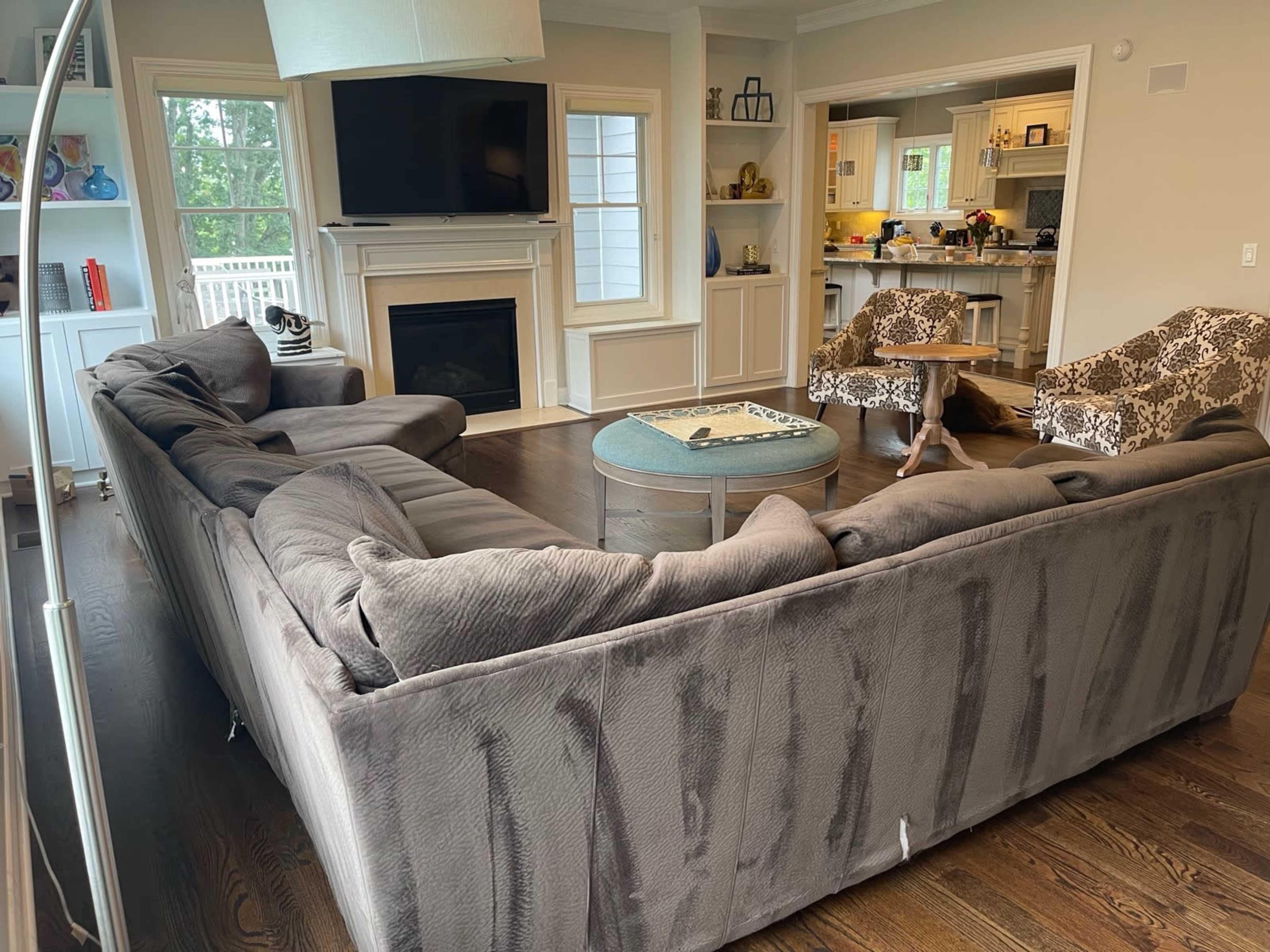 The image shows a spacious living room with a large gray sectional sofa, two patterned chairs, a round coffee table, a fireplace, and a kitchen area visible in the background.