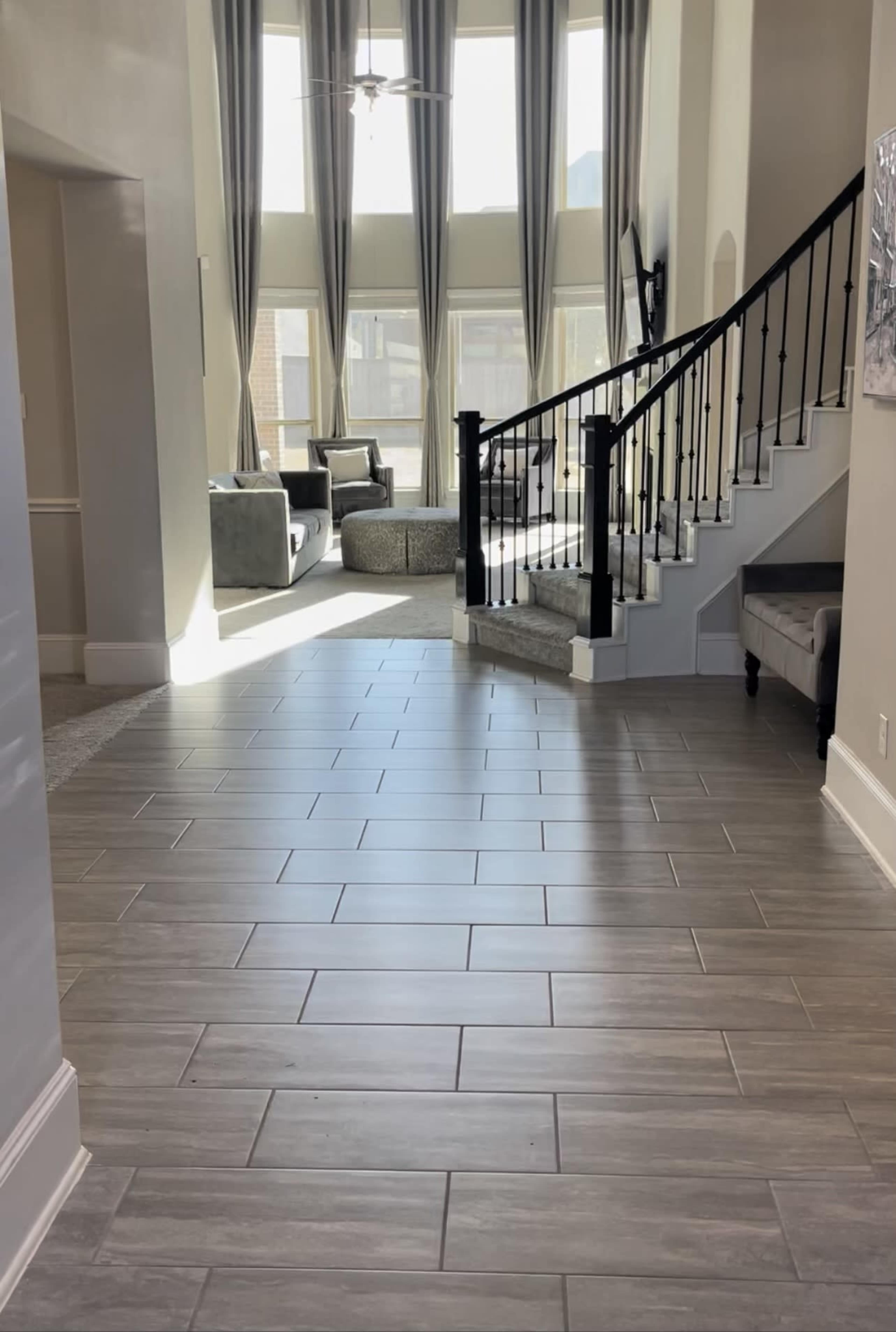 A spacious entryway with a staircase leading to an upper floor, flanked by large windows and a seating area in the background.