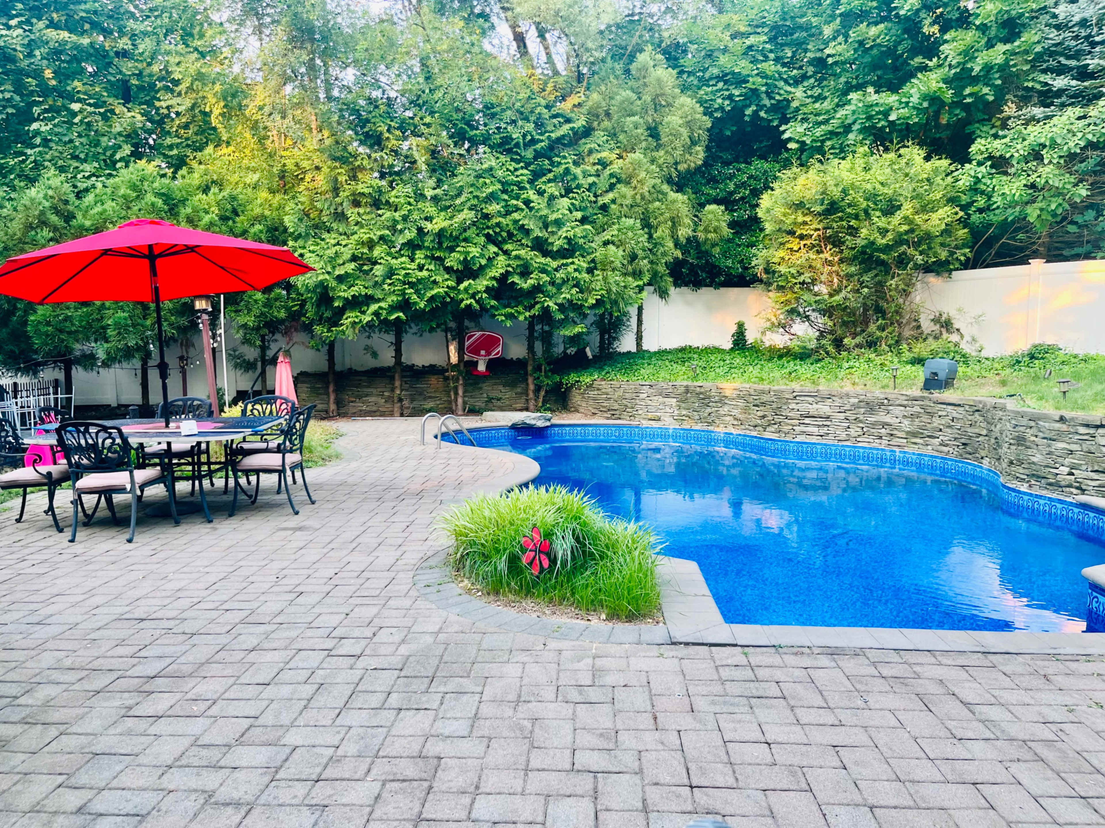 A landscaped backyard features a rectangular swimming pool surrounded by a paved patio with a dining set under a red umbrella.