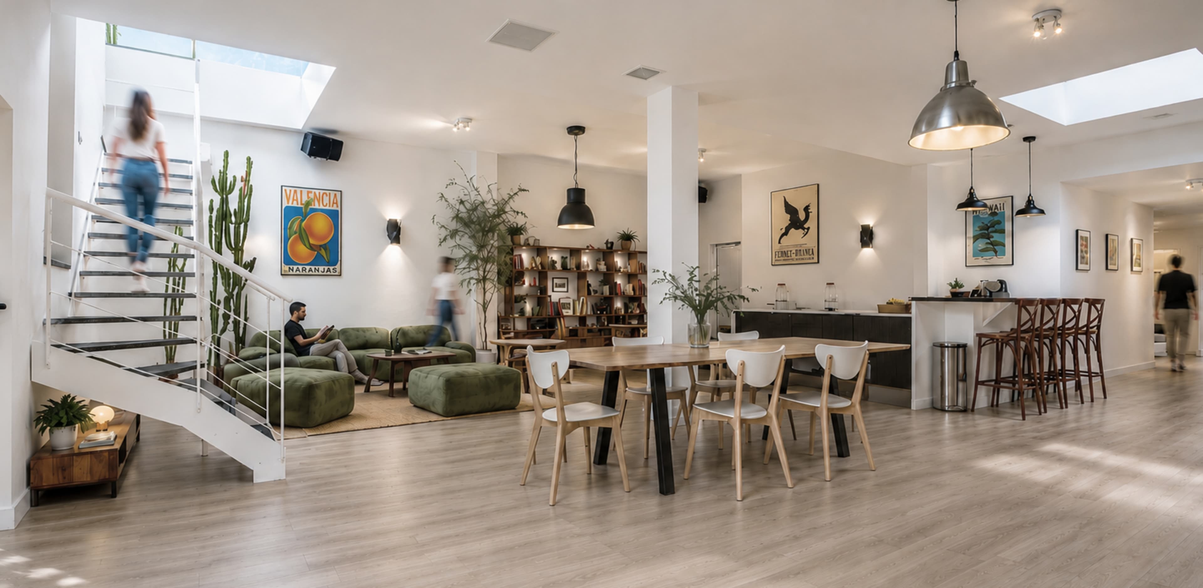 The image shows a modern communal lounge space featuring a combination of green furniture, a dining area with a wooden table and chairs, and decorative plants.