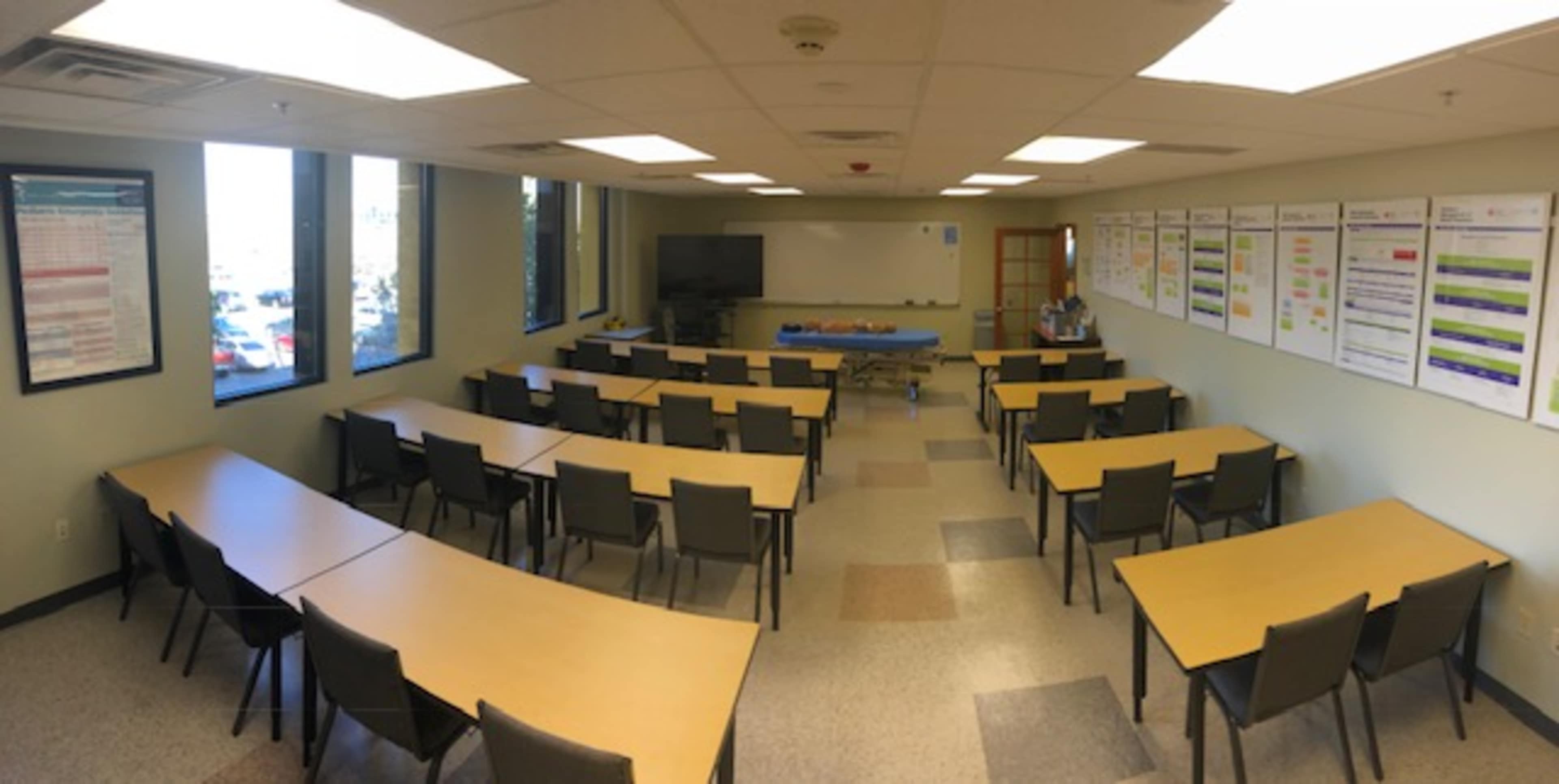 A classroom is set up with several rows of tables and chairs, along with a television and instructional materials displayed on the walls.