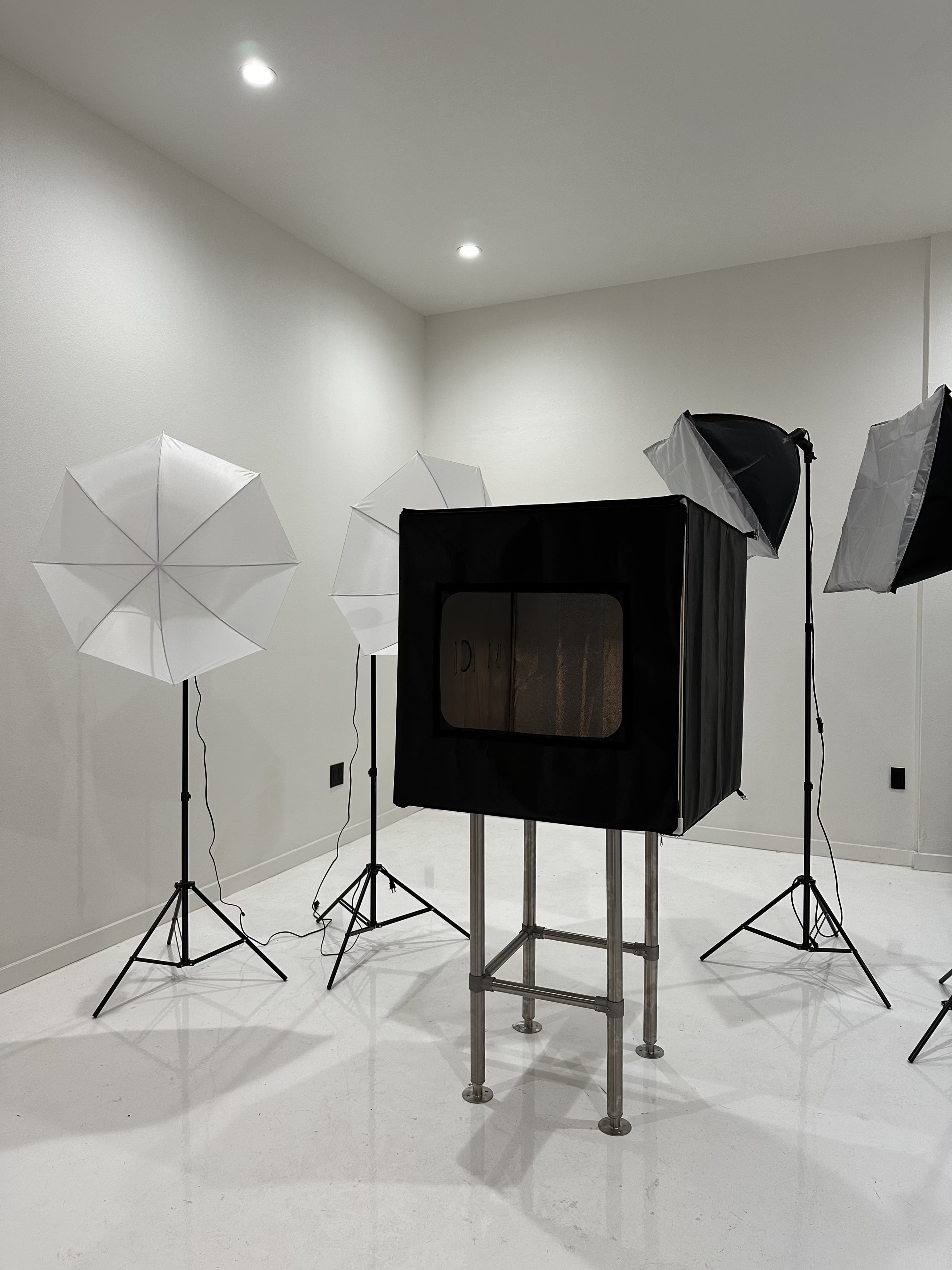 The image depicts a photography studio featuring a black backdrop enclosure surrounded by four light umbrellas and studio lights set on stands.