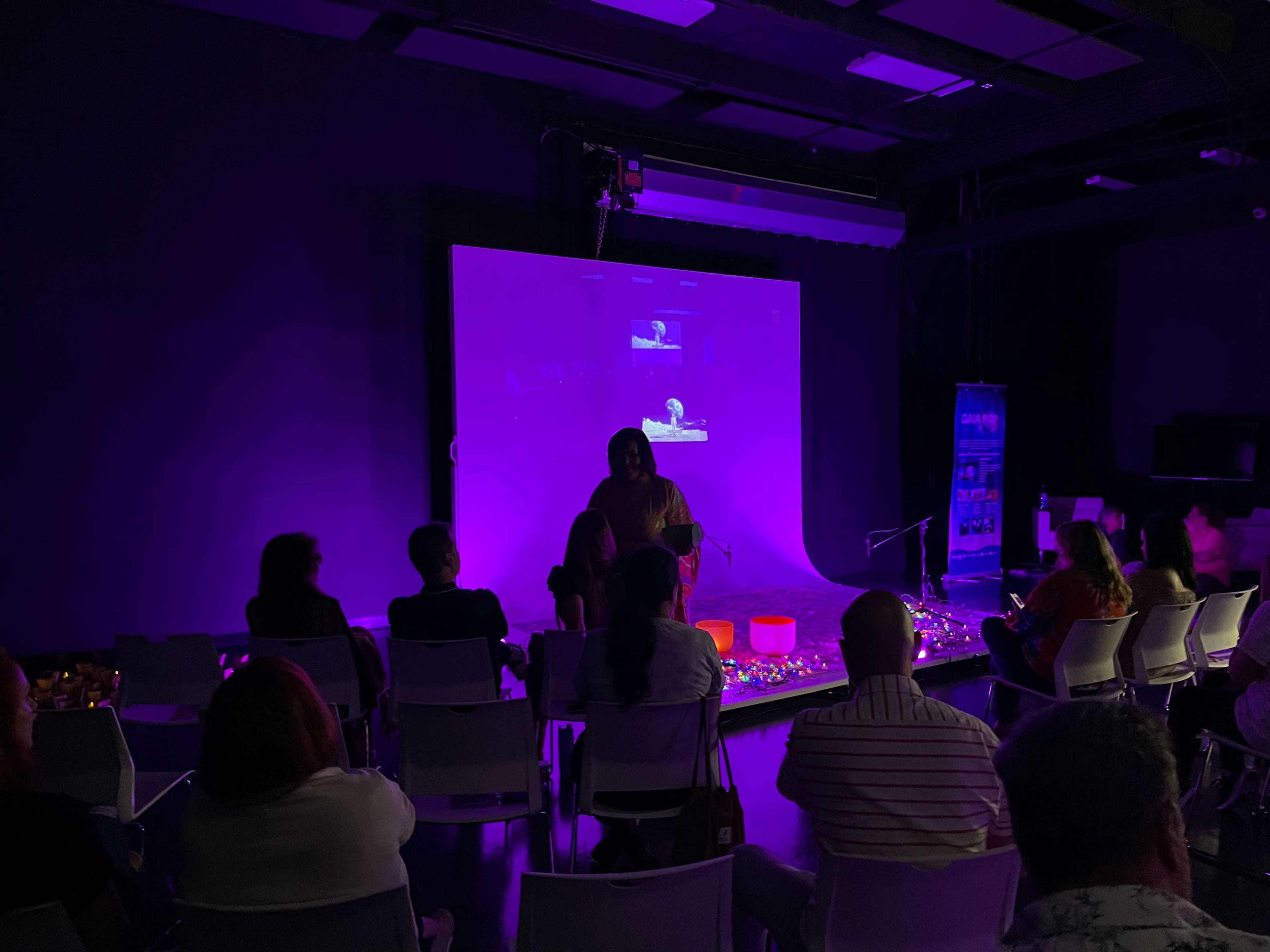 A group of people sits in chairs facing a purple-lit stage where a speaker presents on a large screen.