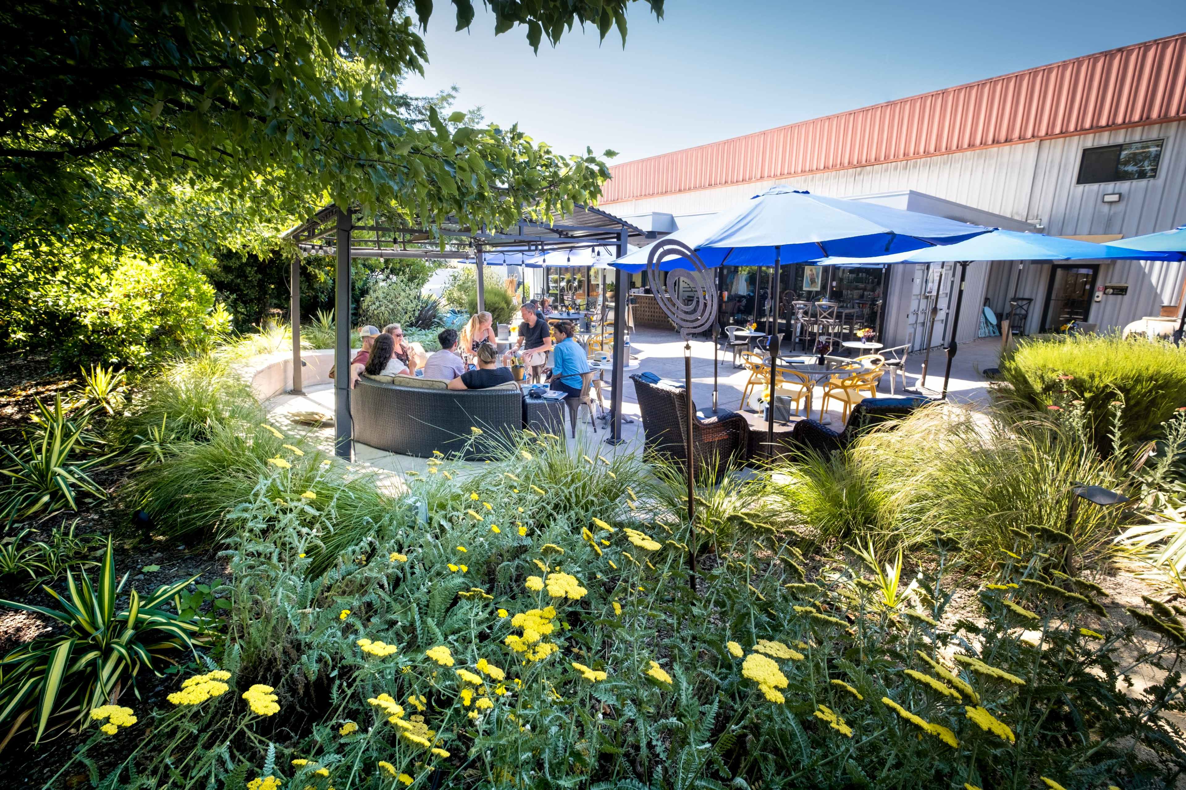 A group of people gathers around a circular seating area under blue umbrellas in a landscaped outdoor space.