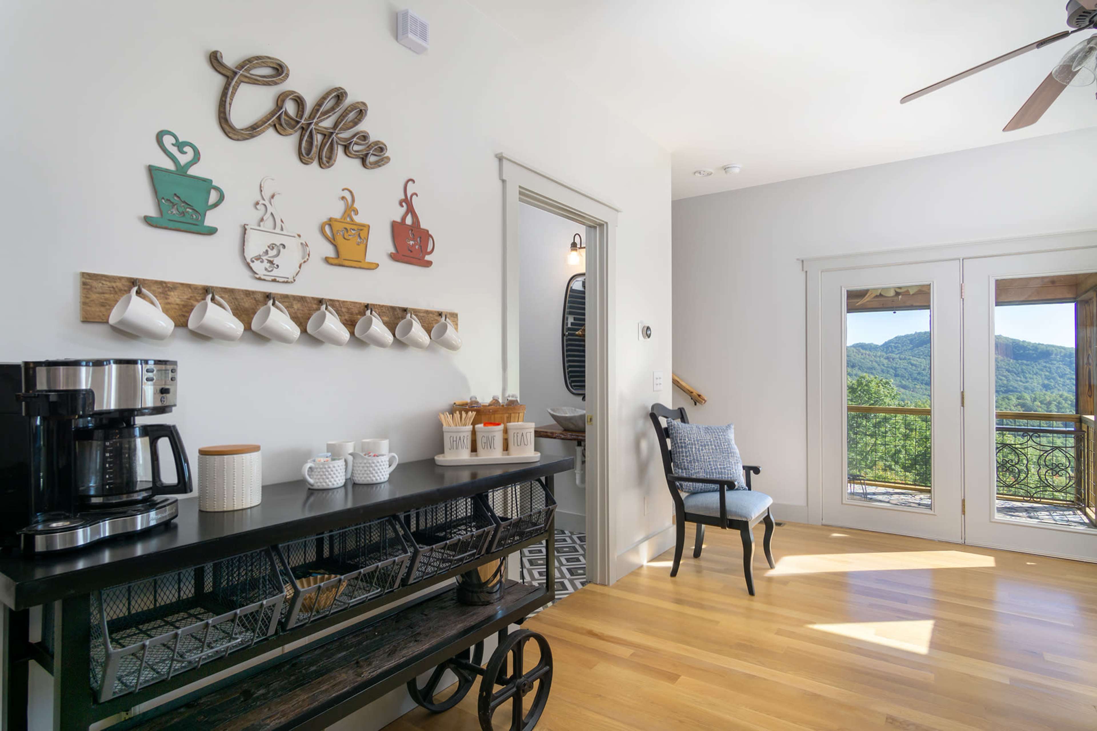 A coffee station featuring a coffee maker, decorative cups, and a sign, is placed against a wall with large windows showcasing a mountain view.