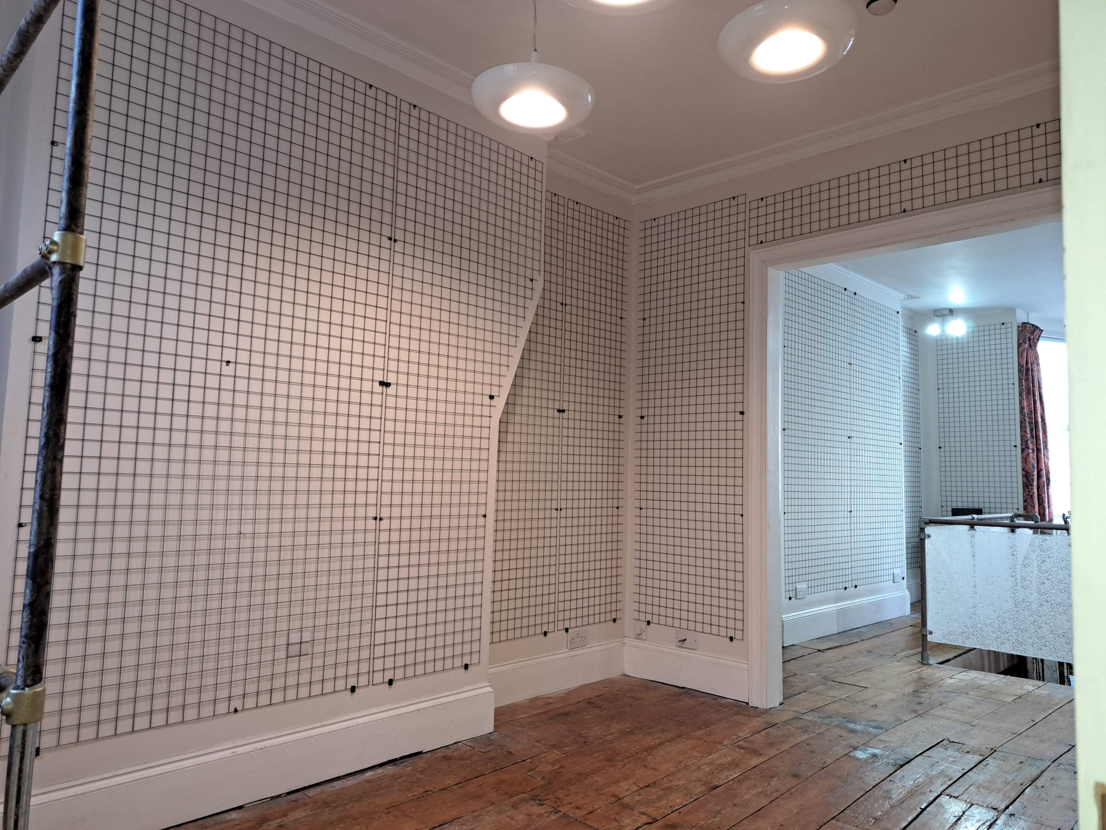An interior room with grid-patterned wall panels and exposed wooden floorboards, illuminated by ceiling lights.