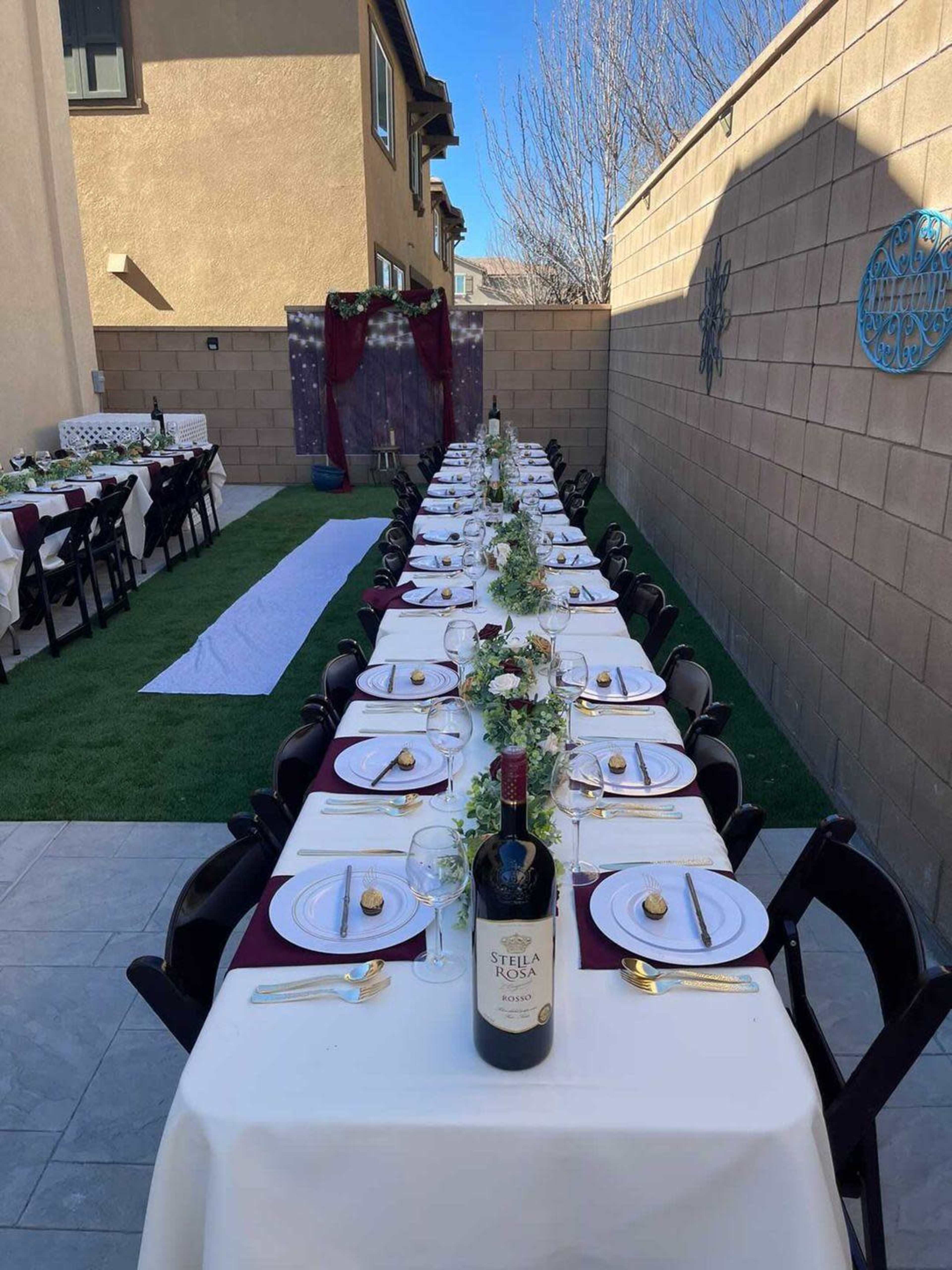 A long, elegantly set dining table with white tablecloths and decorative place settings is arranged outdoors in a narrow backyard.