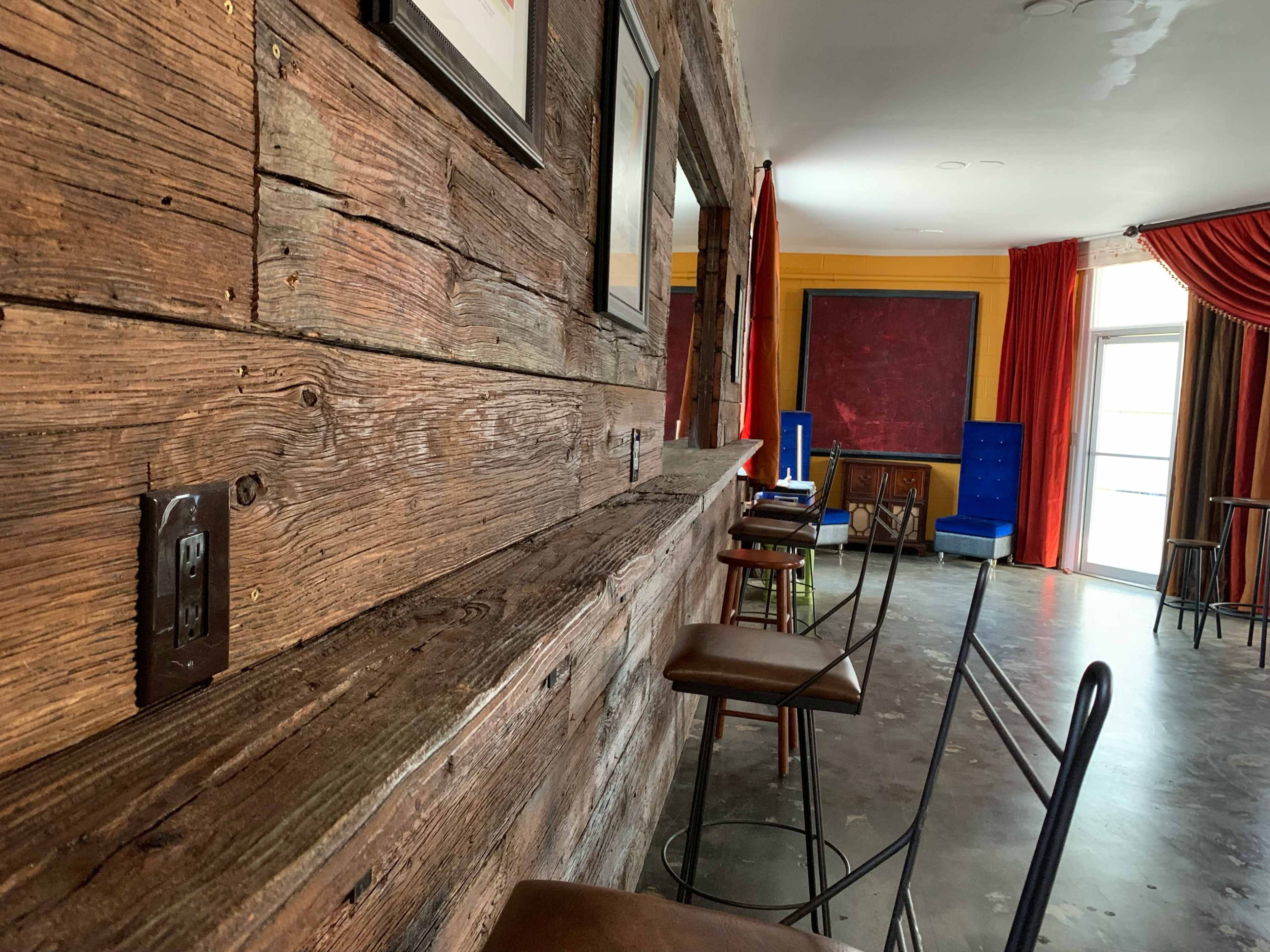 A rustic wooden wall with a bar top, featuring several high stools and a bright room with red drapes and colorful furniture in the background.