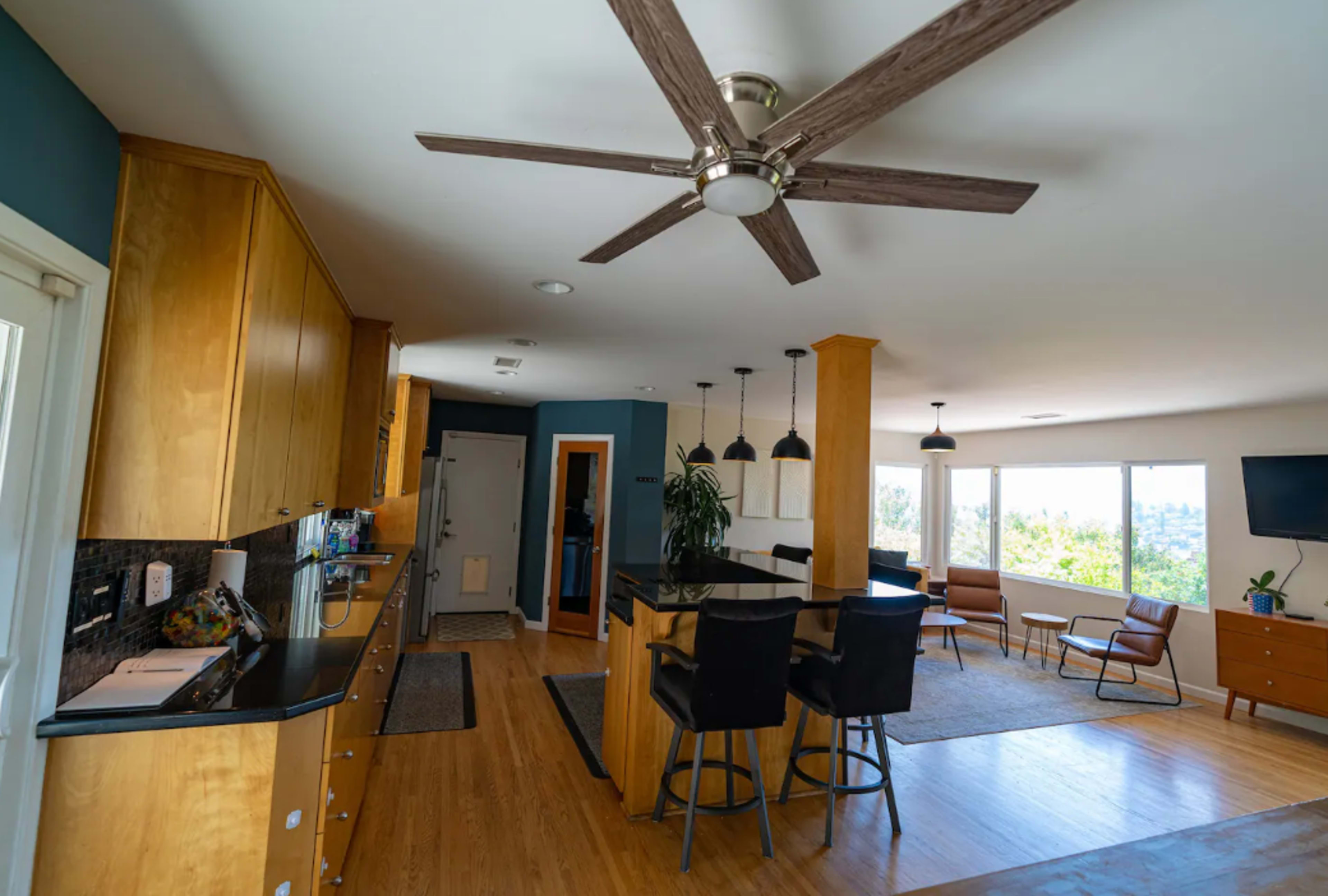 A modern kitchen and living area with wooden cabinets, a large island with bar stools, and a ceiling fan, featuring windows that provide a view of the outdoors.