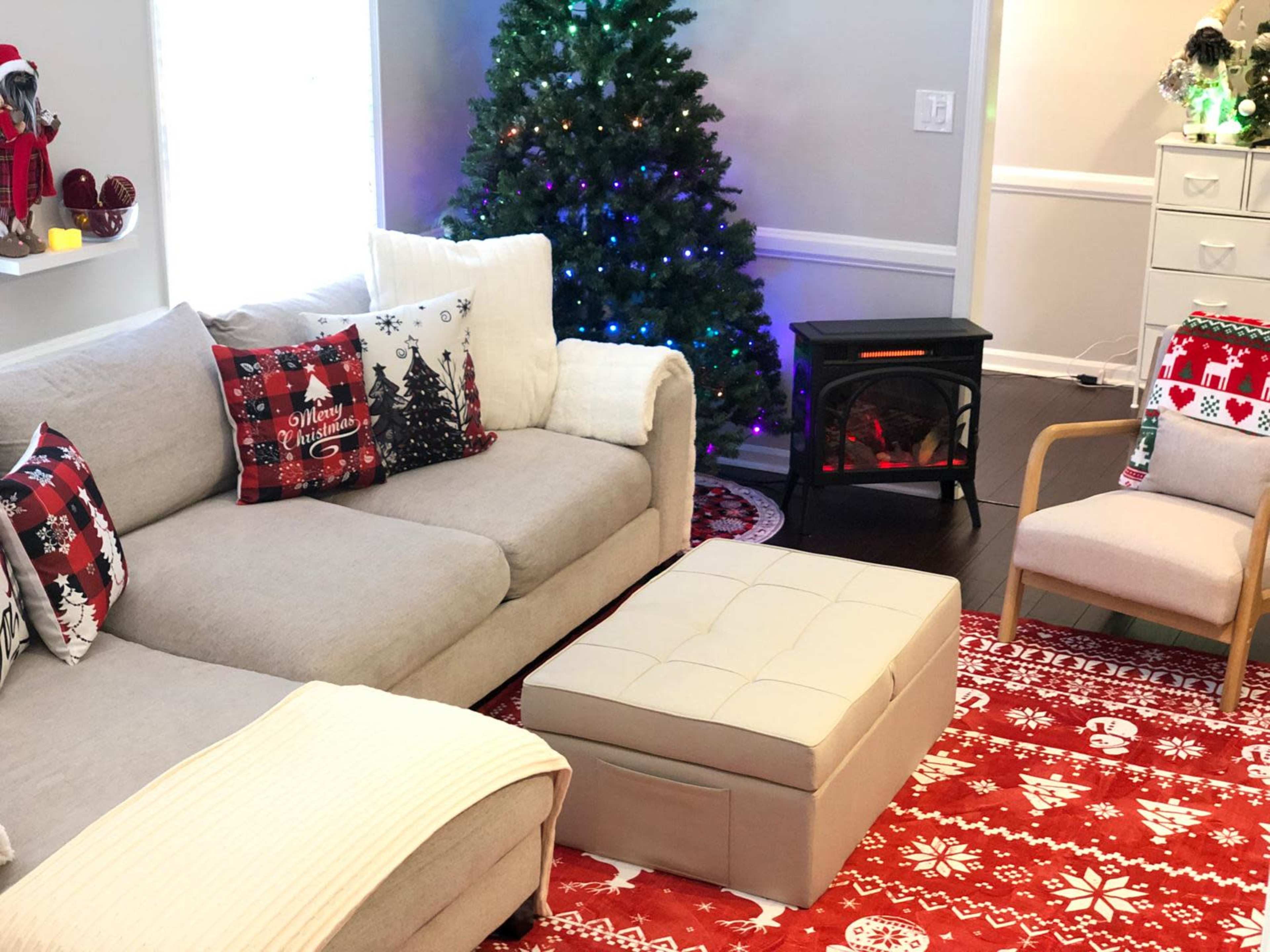 The living room features a gray sectional sofa with decorative pillows, a festive red area rug, a small ottoman, and a cozy electric fireplace beneath a Christmas tree adorned with colorful lights.