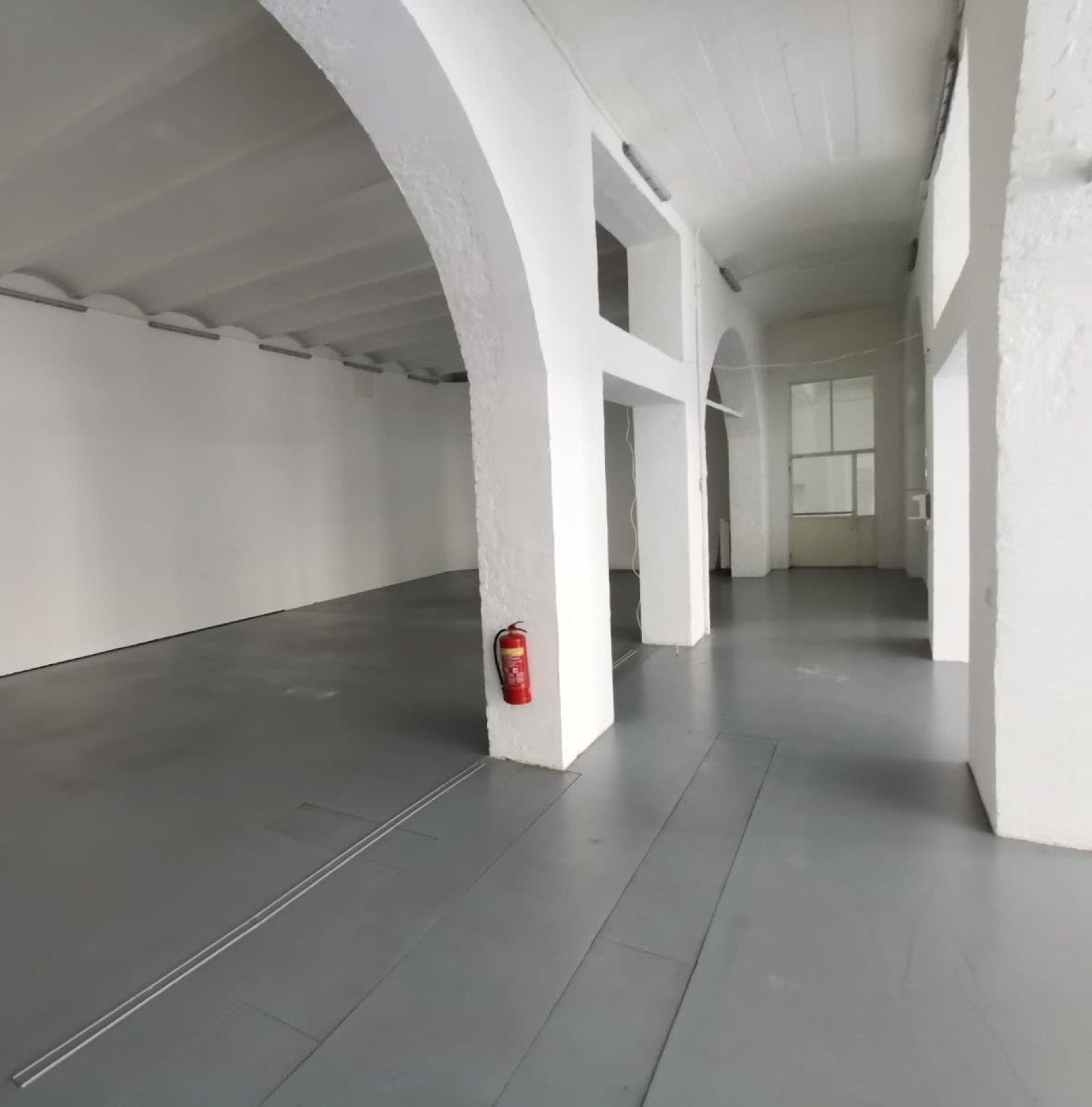 The image shows a spacious, vacant room with high ceilings, white walls, and a gray floor, featuring arched columns and a fire extinguisher on one wall.
