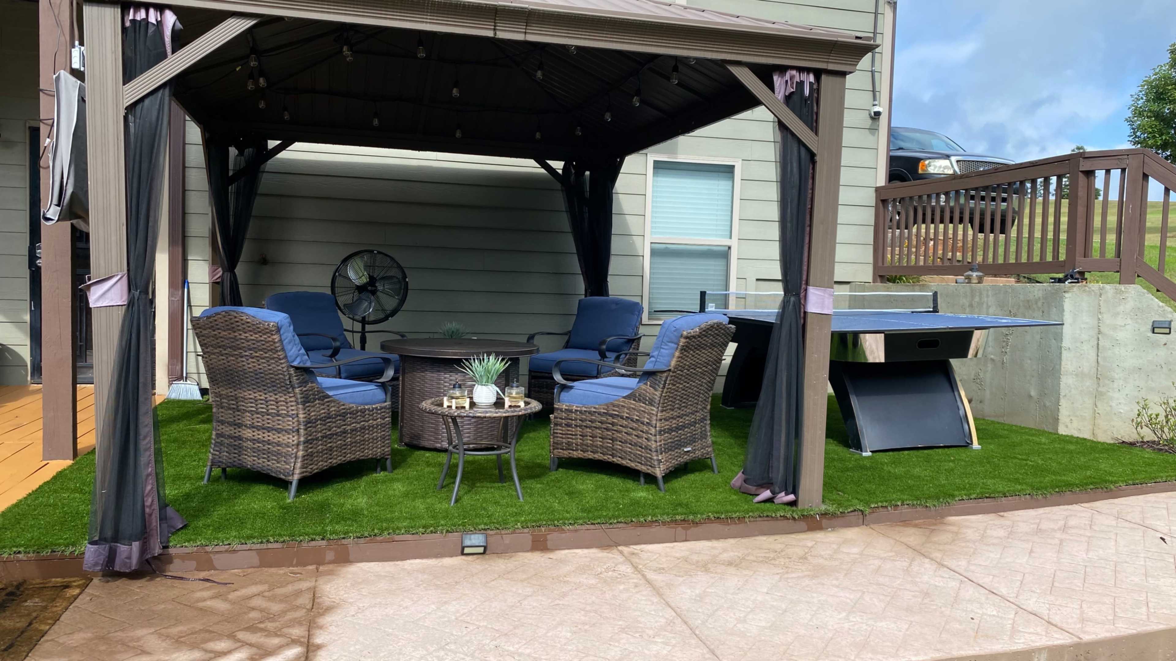 A gazebo with four blue chairs and a table, surrounded by artificial grass, and a ping pong table nearby.