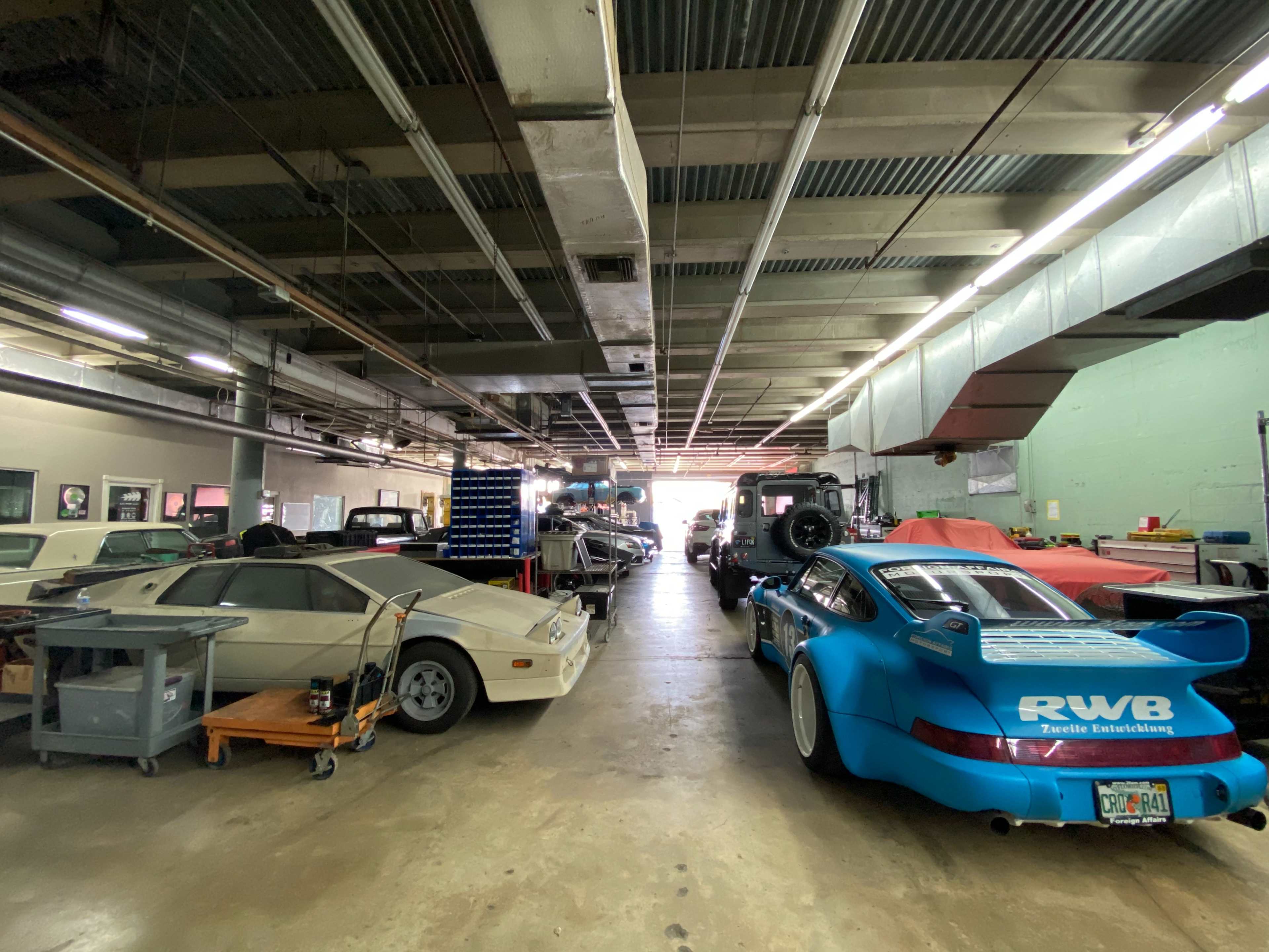 The image shows an automotive workshop with various cars, including a blue racing Porsche, lined up along either side of a wide aisle.