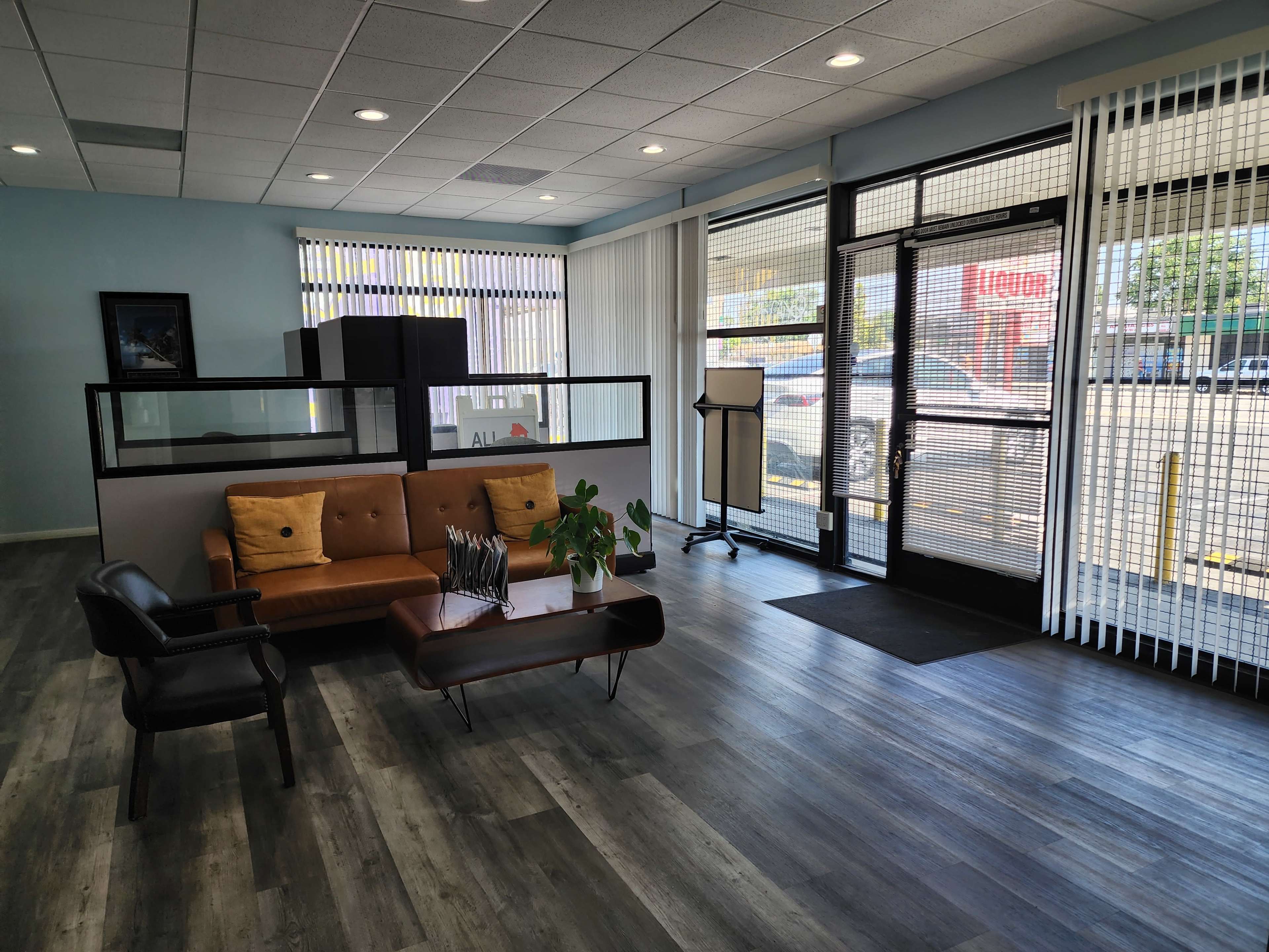 The image shows a modern waiting area with a brown leather couch, a coffee table, and a large front window facing a parking lot.