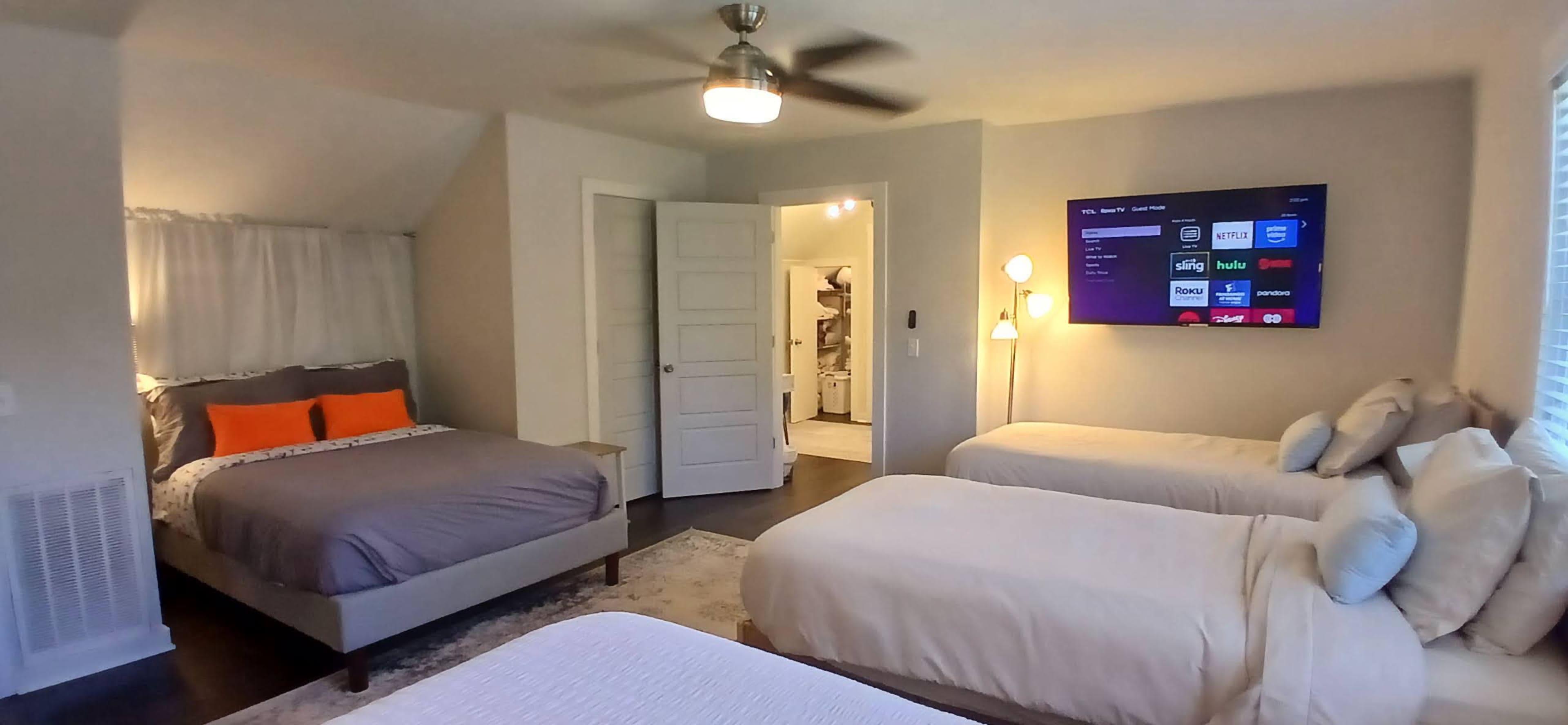 The image shows a well-lit bedroom featuring three beds, a television mounted on the wall, and a door leading to a bathroom area.