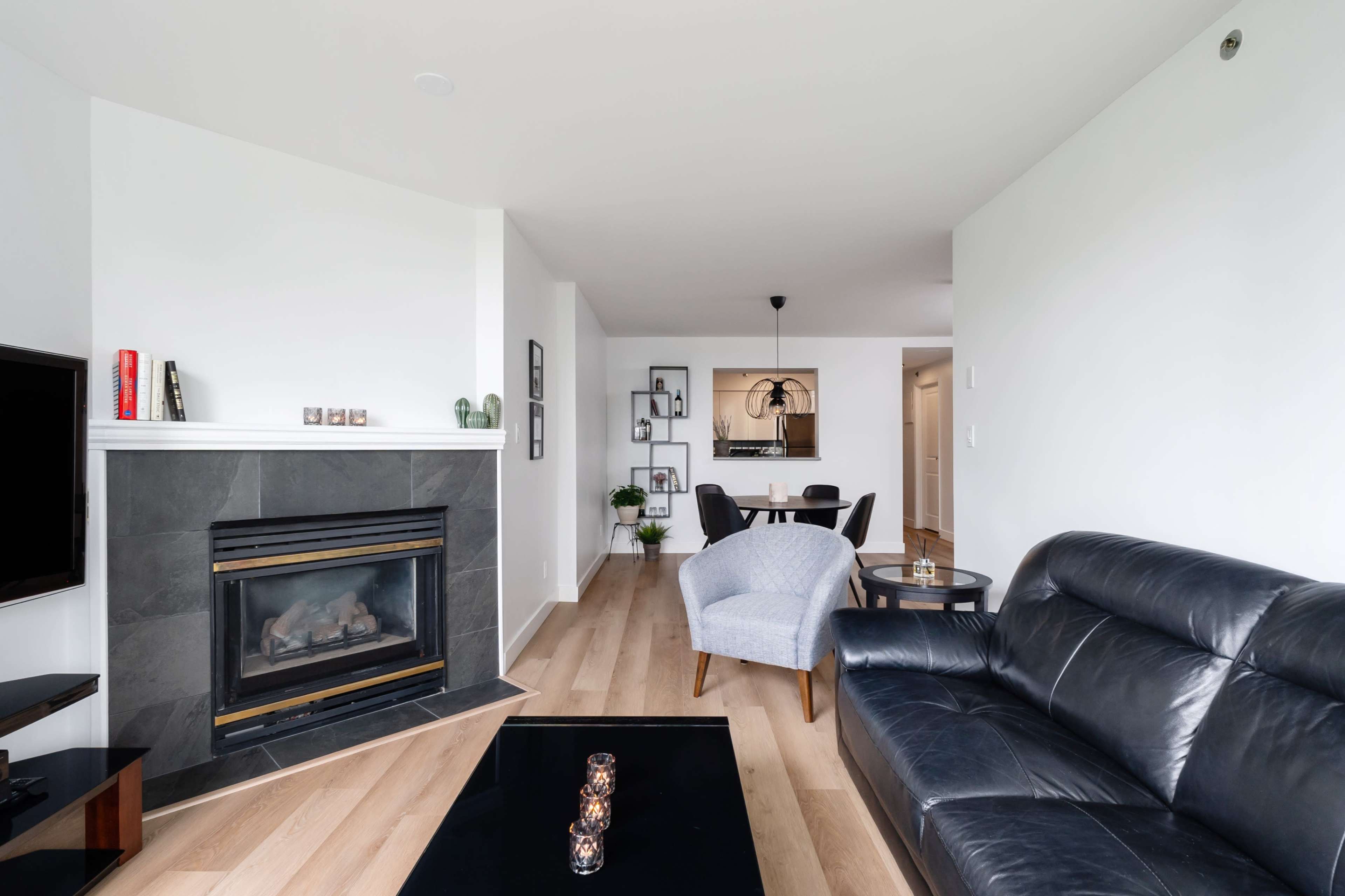 A modern living room featuring a black leather sofa, a sleek coffee table, a fireplace with a stone surround, and a dining area in the background.