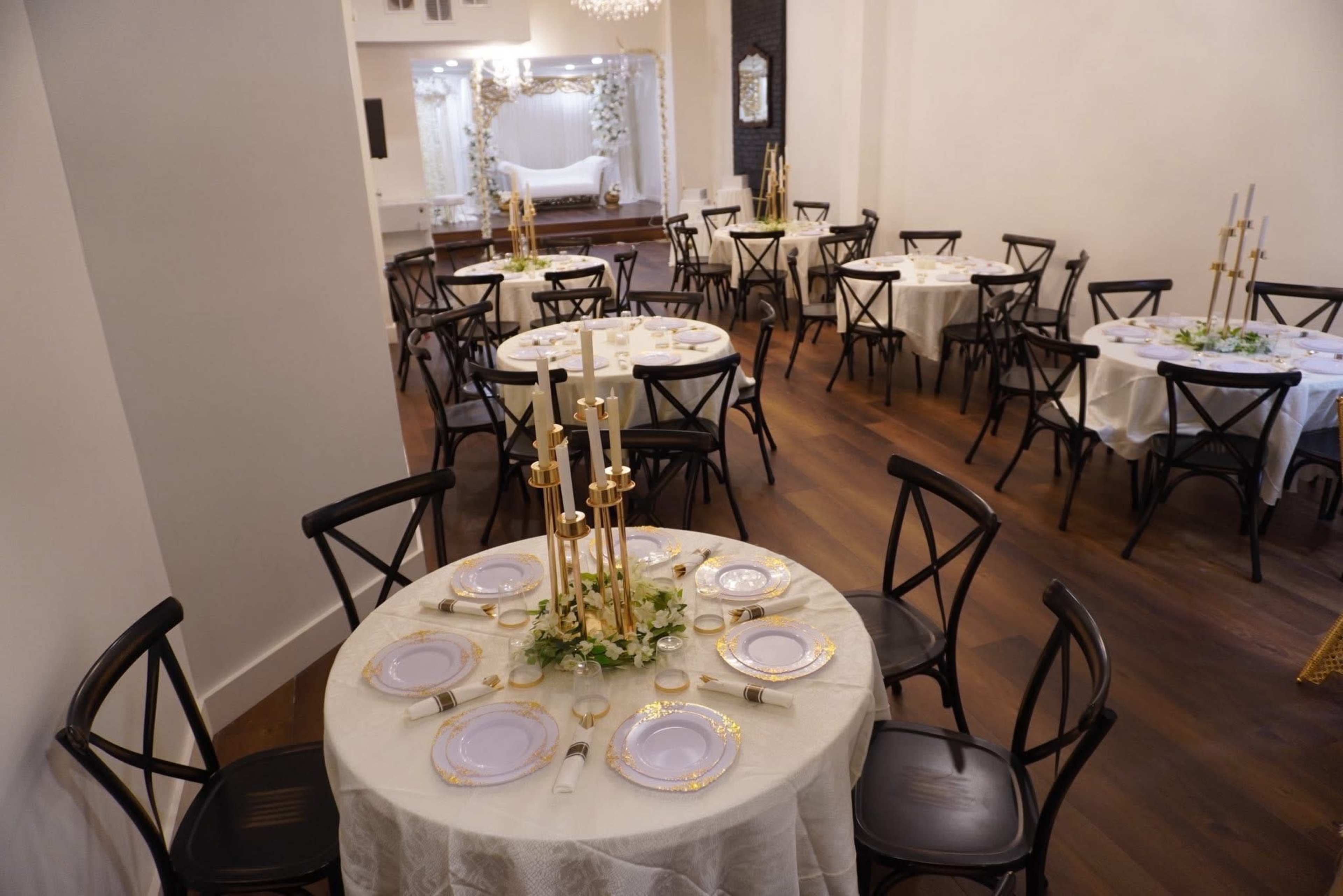 The image shows a neatly arranged dining area with several round tables set for a formal event, featuring elegant white tablecloths, plates, and tall candle holders.