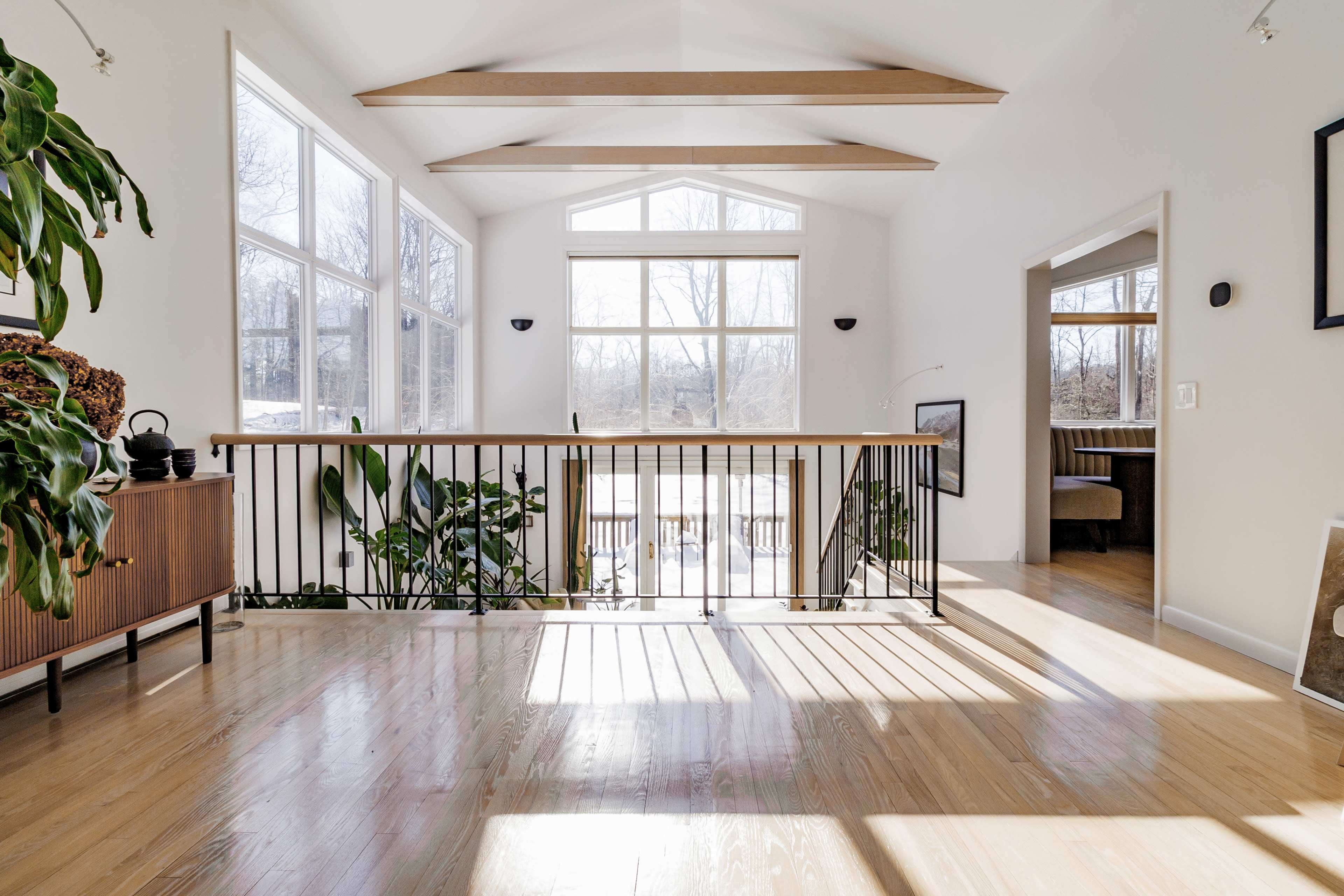 A bright, open room features large windows, wooden beams, and plants, with a balcony overlooking a sunlit area.