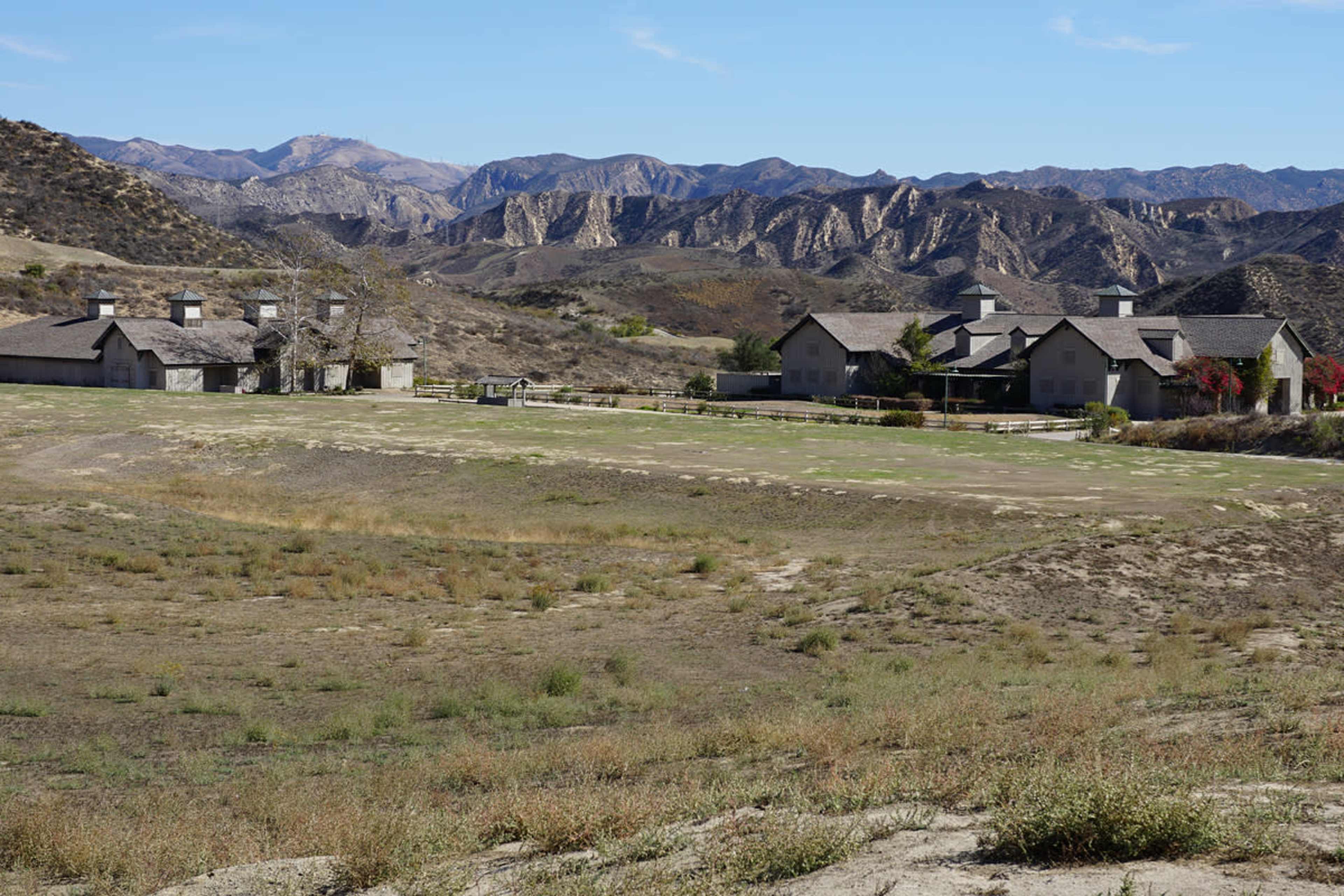 The image shows a rural landscape with several houses nestled near a dry, grassy area and mountains in the background.