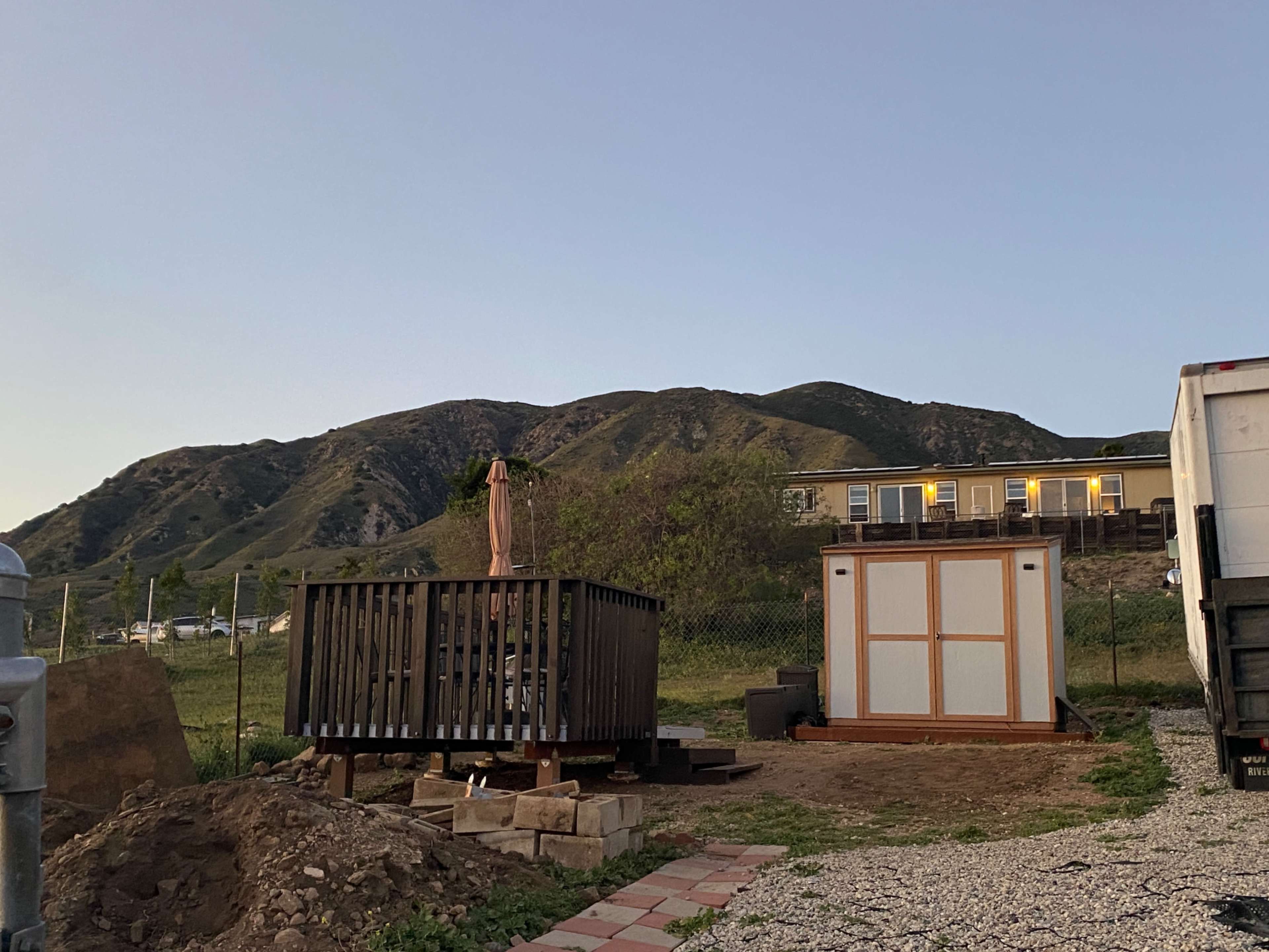 A wooden deck with an umbrella is shown next to a storage shed in a rural area with a hillside in the background.