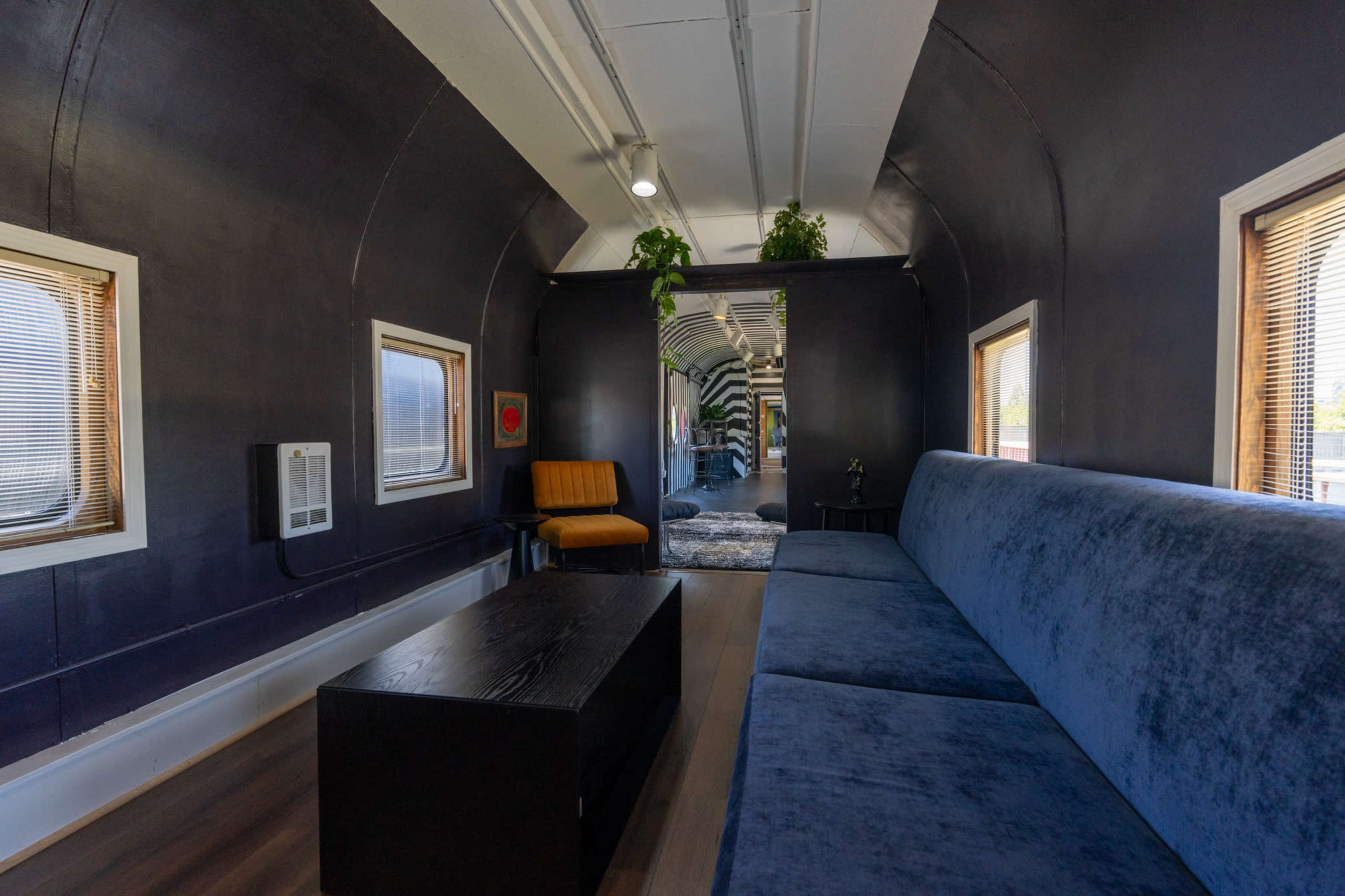 The image shows the interior of a train car converted into a living space, featuring a blue couch, a wooden coffee table, and large windows with blinds.