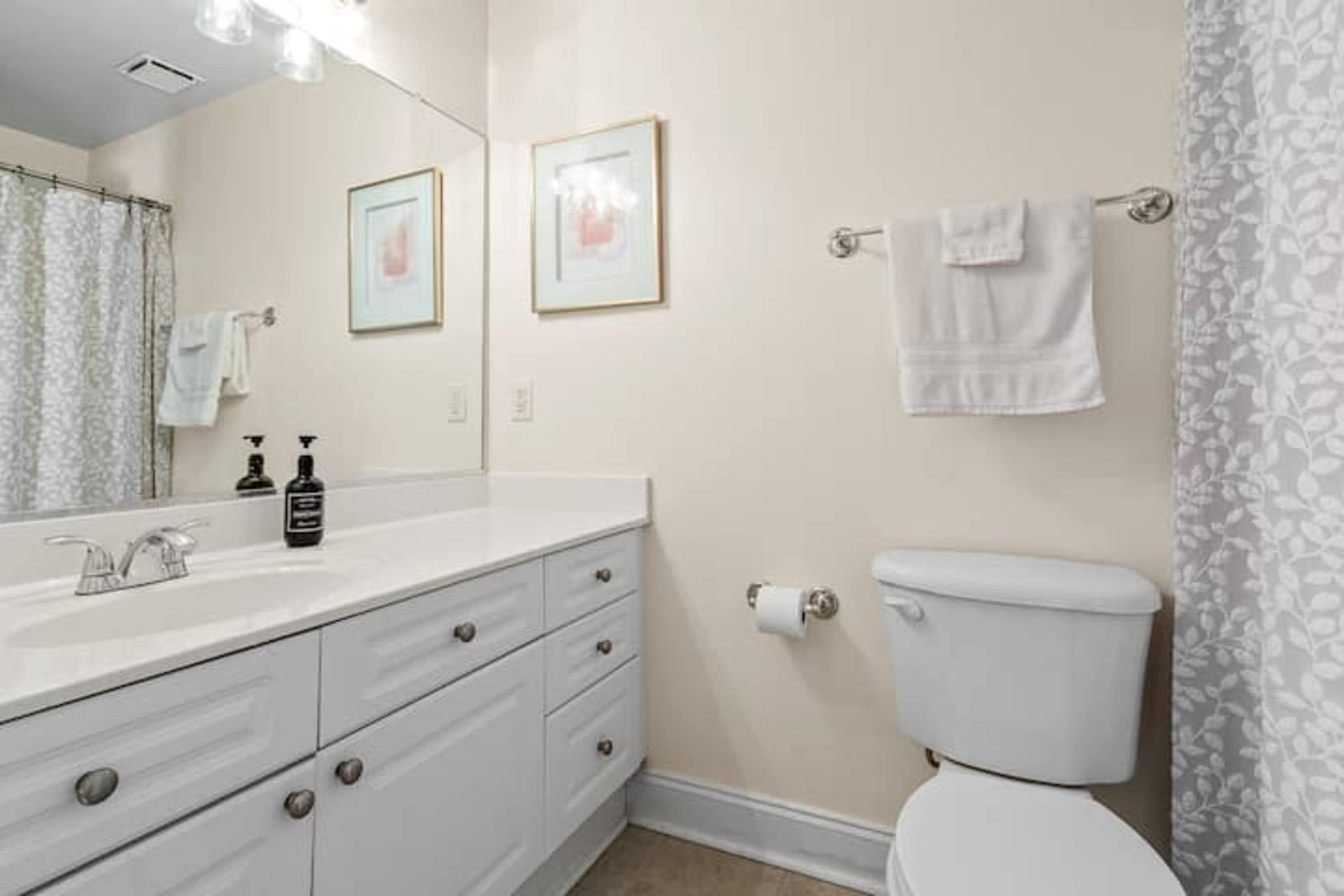 The image shows a bathroom with a white vanity, a large mirror, a towel rack with a towel, and a shower curtain with a floral pattern.