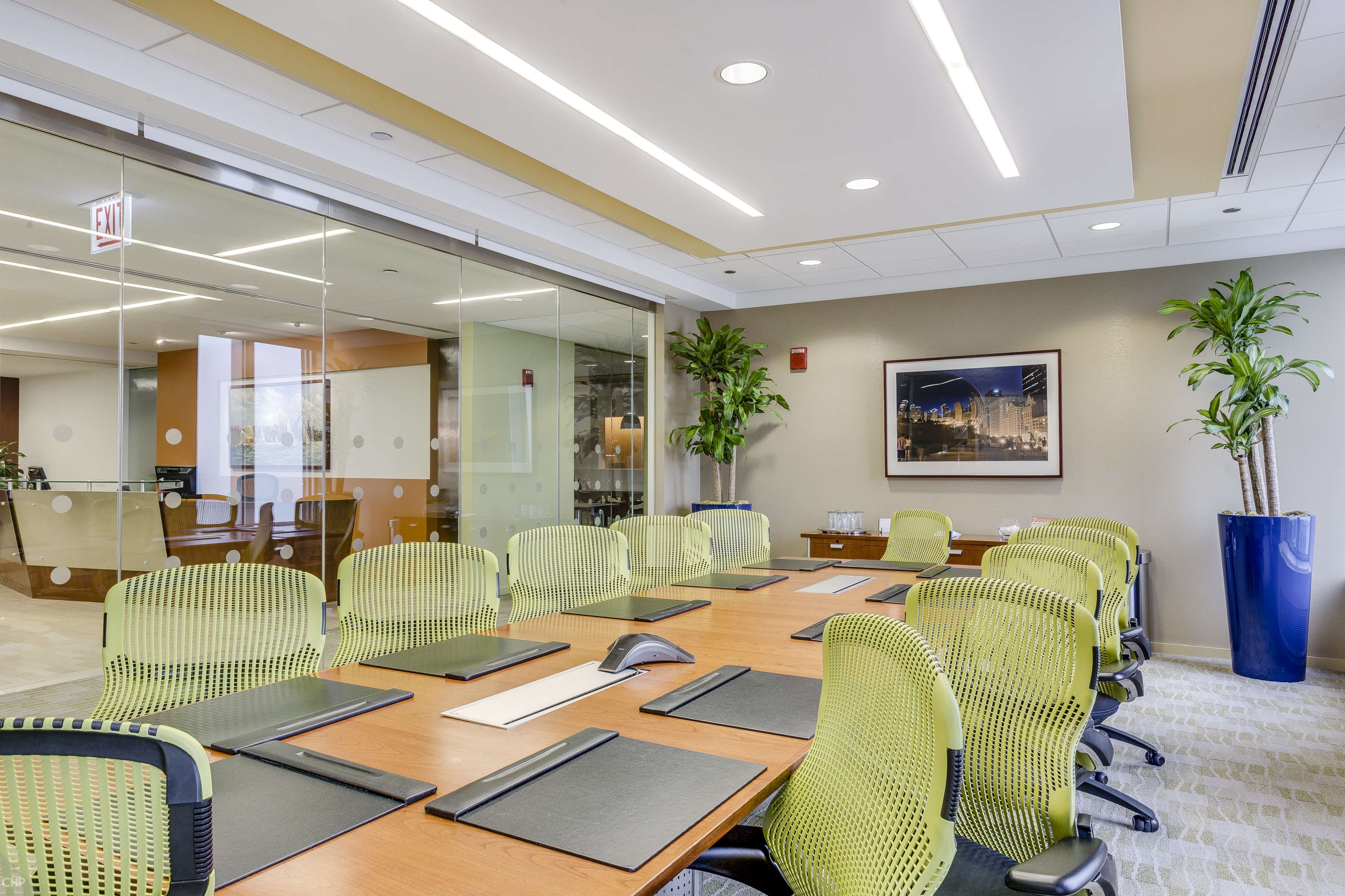 A modern conference room features a large wooden table surrounded by green chairs, with glass walls and potted plants.