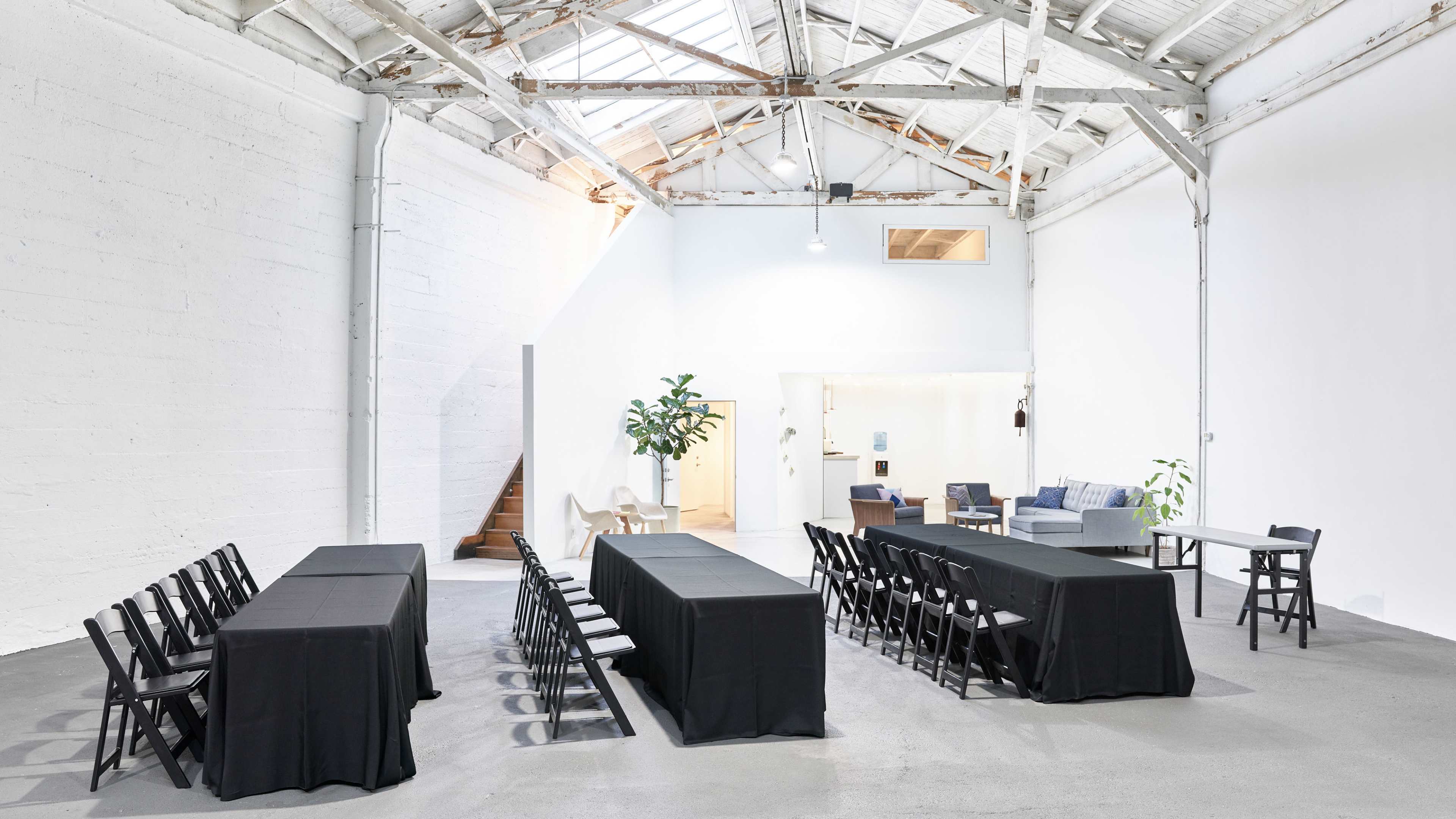 A spacious room features two rows of long tables covered with black tablecloths, surrounded by folding chairs, under a high ceiling with skylights.
