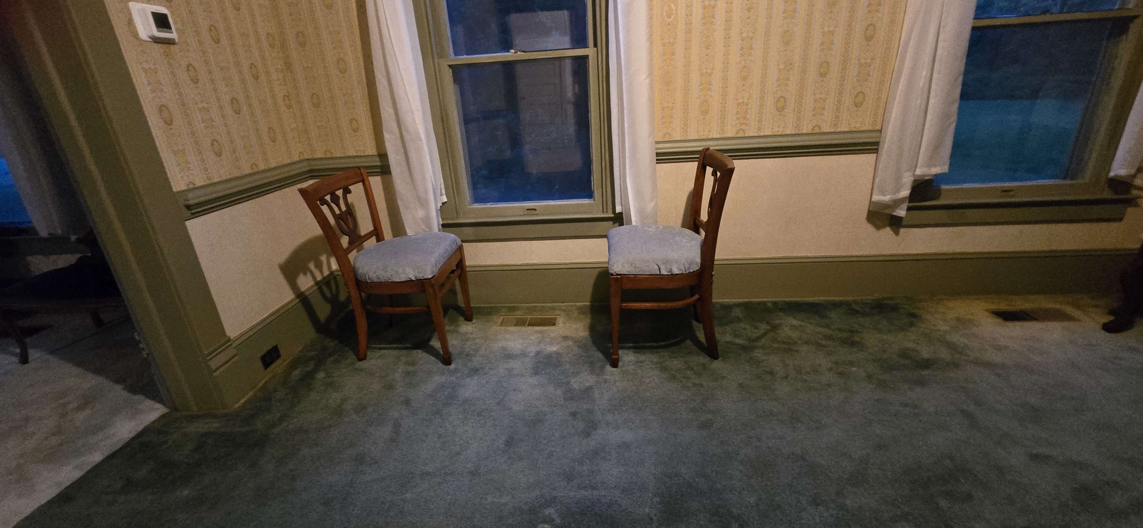 The image shows a room with two wooden chairs placed in a corner, on a green carpet with wallpapered walls.