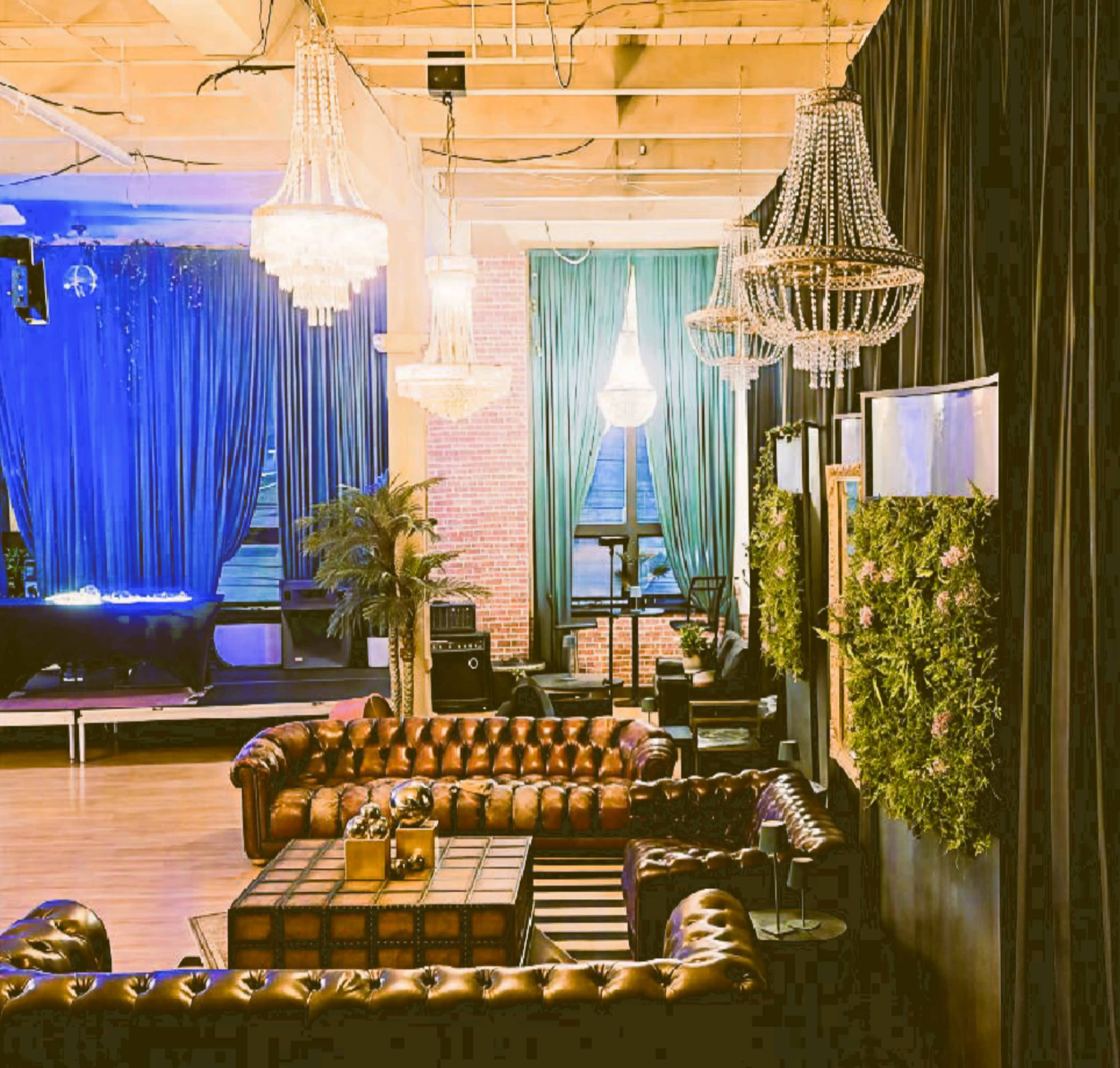 The image shows a stylish indoor lounge area with leather sofas, ornate chandeliers, and decorative greenery against a backdrop of rich curtains and wooden beams.